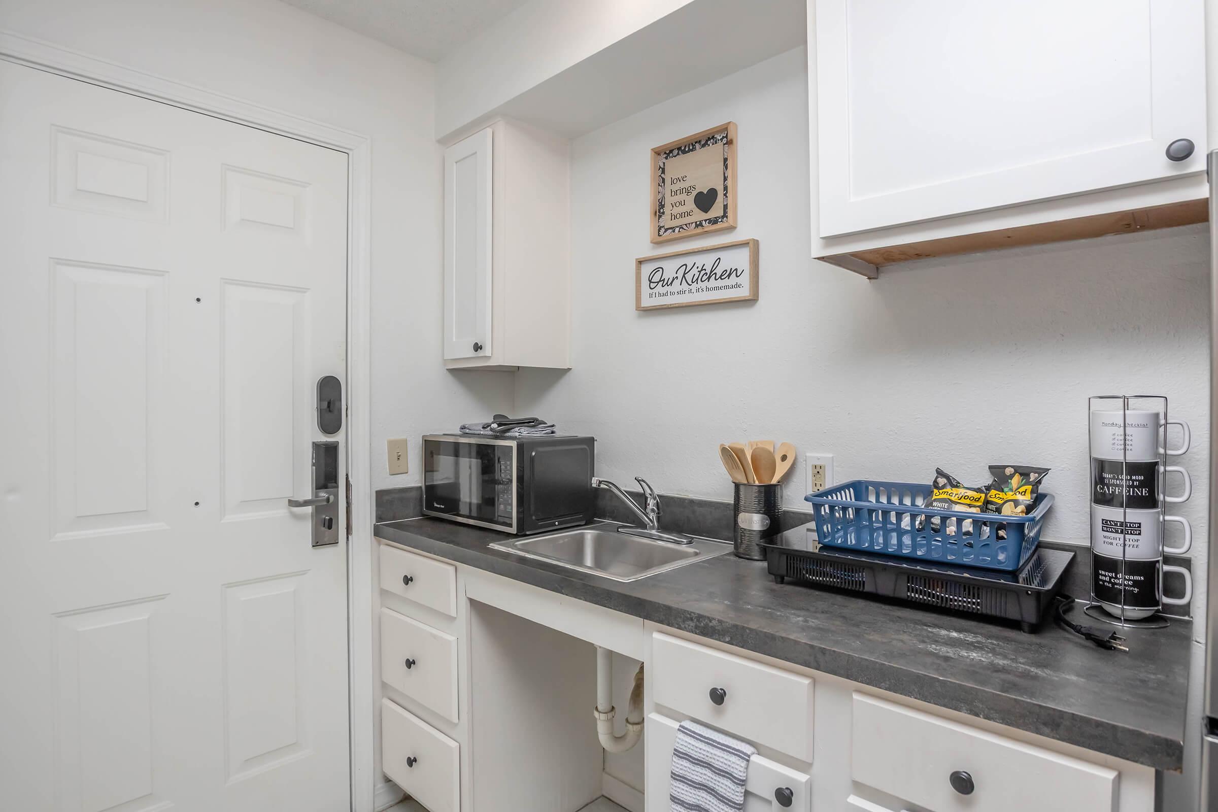 A small kitchen space featuring a door, a sink, a microwave, and a countertop with utensils. A blue dish rack sits beside the sink, and there are white cabinets above with a decorative sign that reads "Be-Kitchen." The overall aesthetic is clean and simple, with neutral colors.