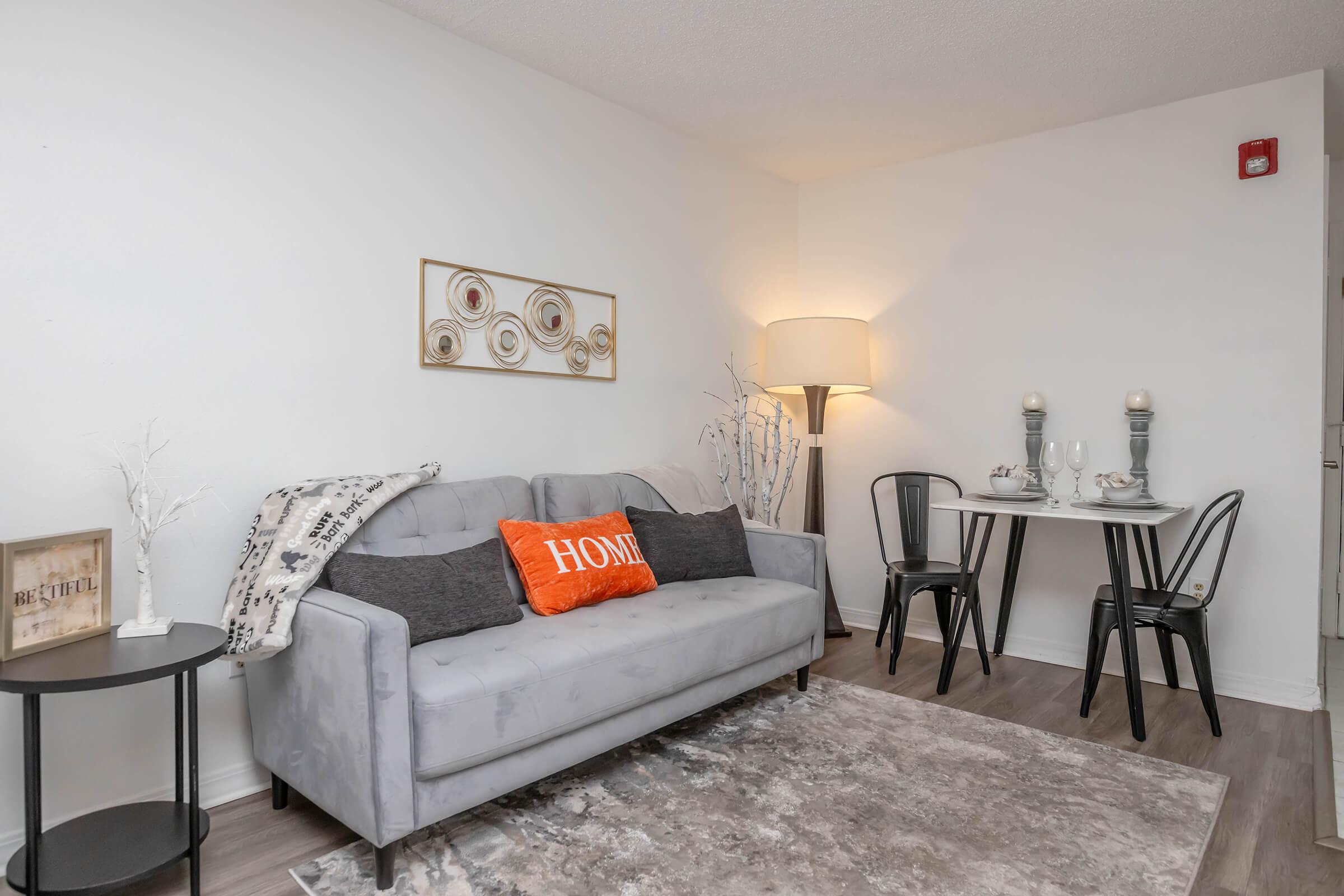 A cozy living area featuring a light gray sofa adorned with decorative pillows, including an orange one that reads "HOME." A small dining table with black chairs, set for two, is positioned nearby. The space is bright and minimalistic, with a modern lamp and wall decor adding character.