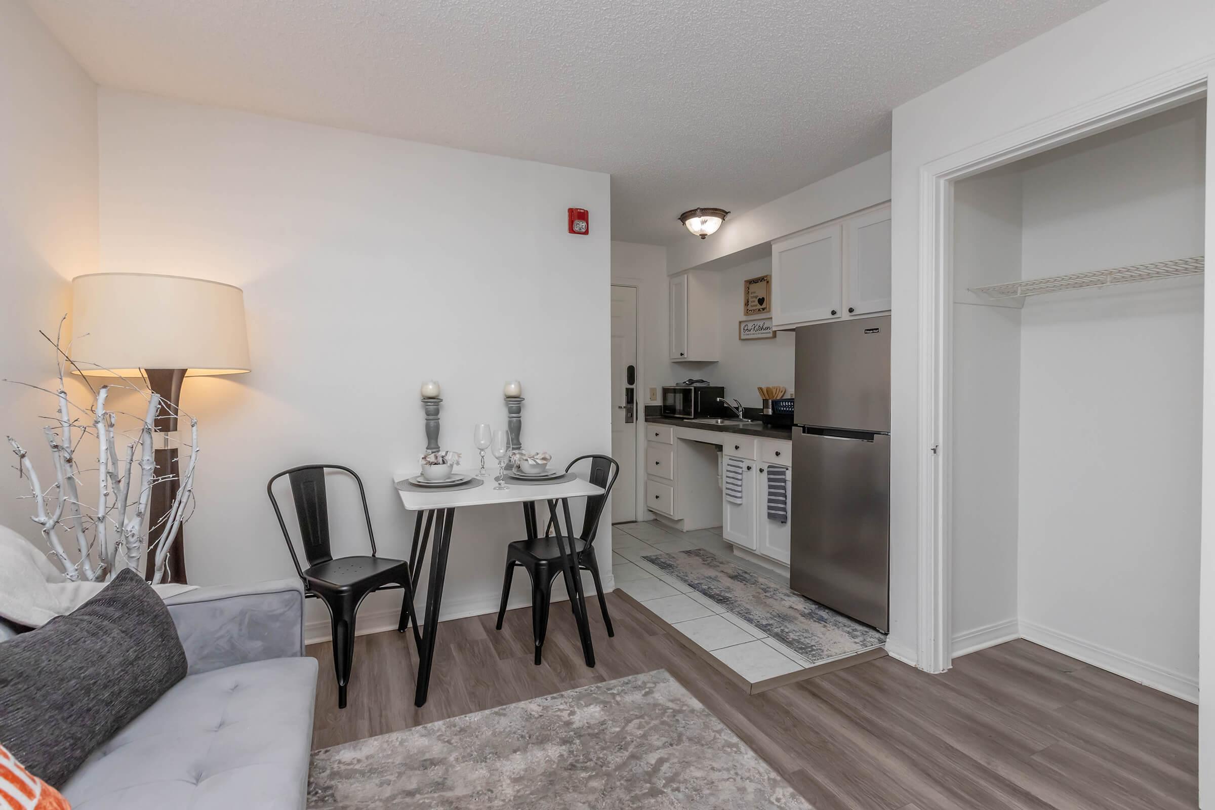 A modern, cozy apartment interior featuring a small dining table set for two, a comfortable gray sofa, a lamp, and a kitchenette with stainless steel appliances. The flooring is light wood, and there is a small closet space visible. The overall decor is minimalist and inviting.