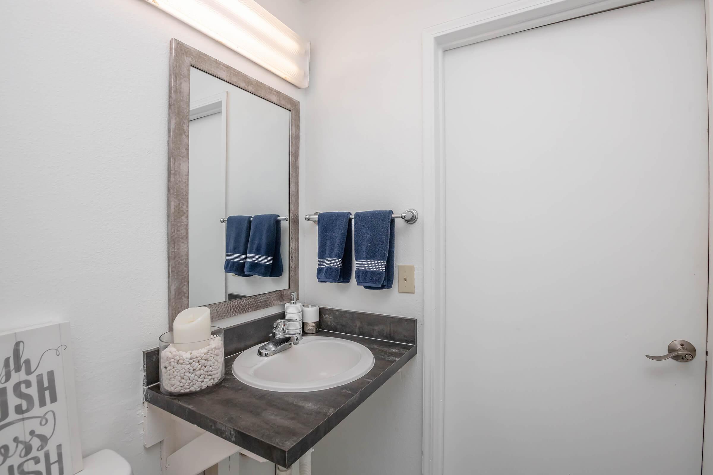 A clean and tidy bathroom featuring a round sink with a gray countertop, a large mirror above, a soap dispenser, and decorative items. Two blue towels hang on a towel rack. The wall is painted white, and a door leads to the adjoining space. A decorative jar filled with small items is on the countertop.