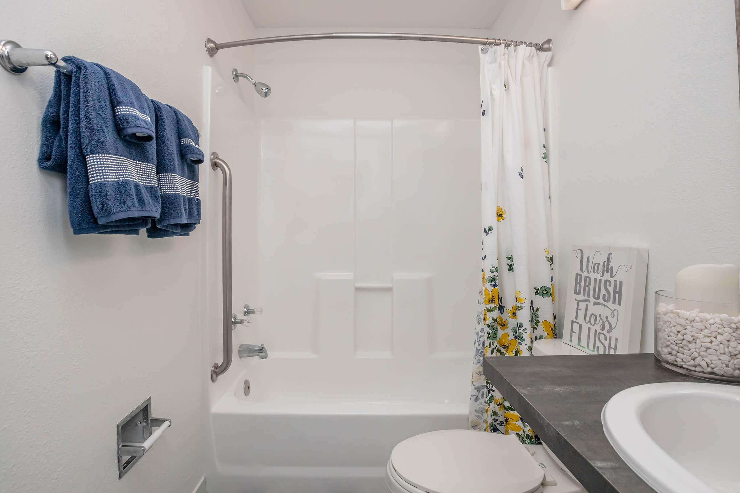 A clean, modern bathroom featuring a white bathtub with a shower curtain decorated with flowers. There are blue towels hanging on a rack, and a decorative sign that reads "Brush, Flush, Flush." The countertop includes a container with white pebbles, enhancing the serene atmosphere.