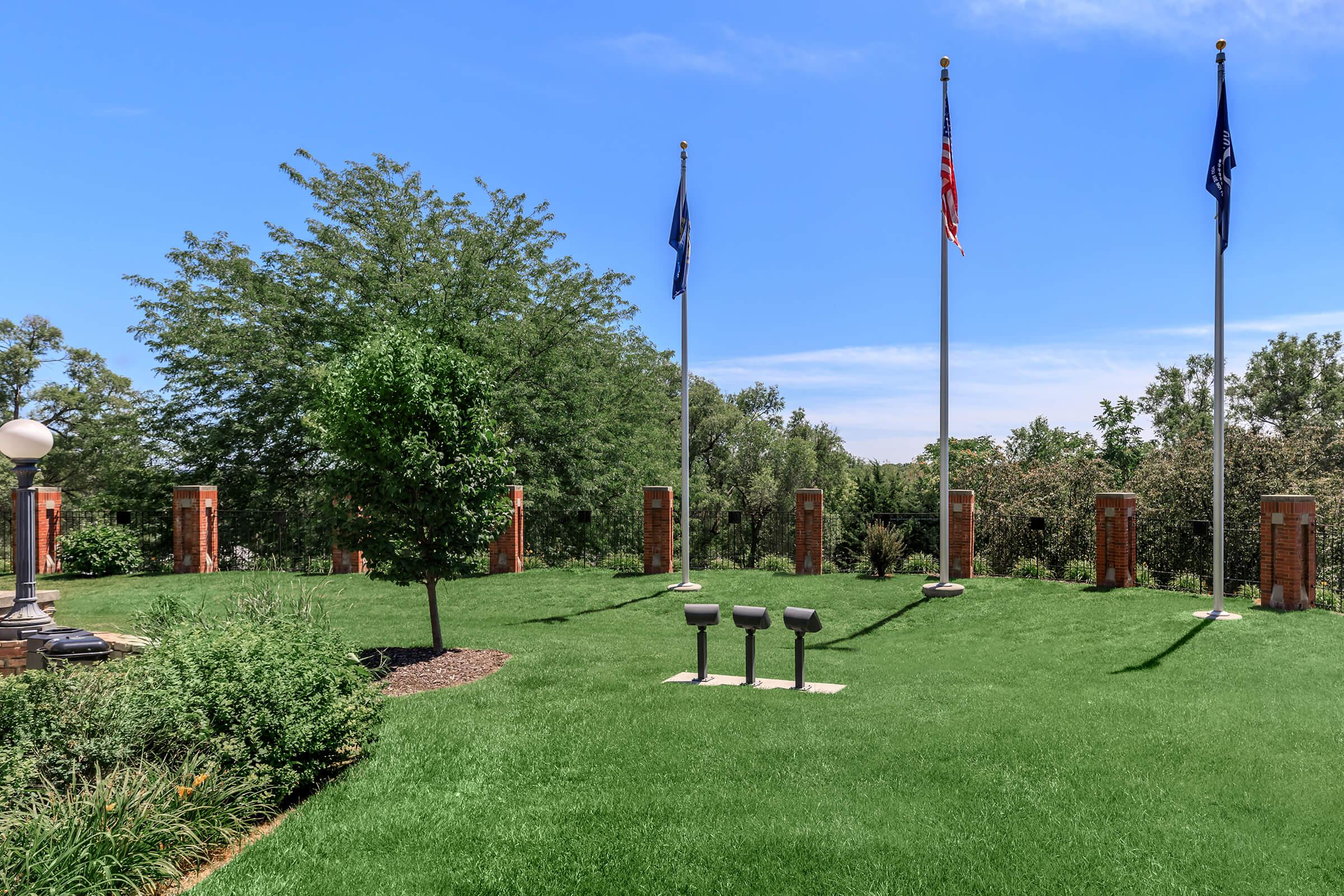 A grassy area with three flagpoles displaying flags, surrounded by greenery and trees. There are decorative brick pillars in the background and a small light fixture to the left. The sky is clear and blue, indicating a sunny day.