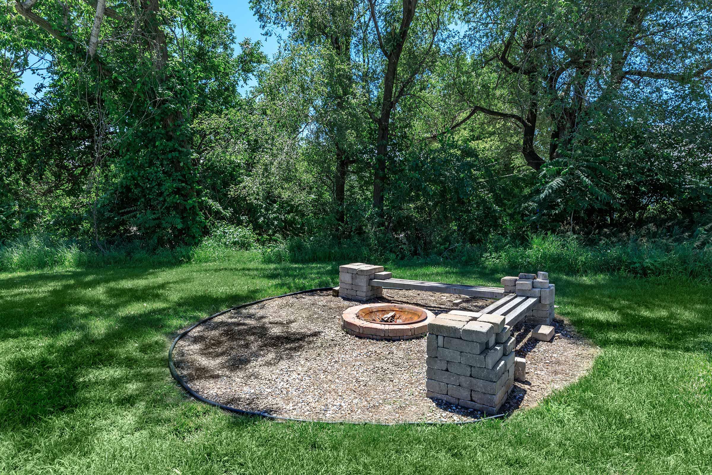 A circular fire pit surrounded by stone seating in a grassy clearing, with trees and lush greenery in the background under a clear blue sky. The scene is peaceful and suitable for outdoor gatherings.