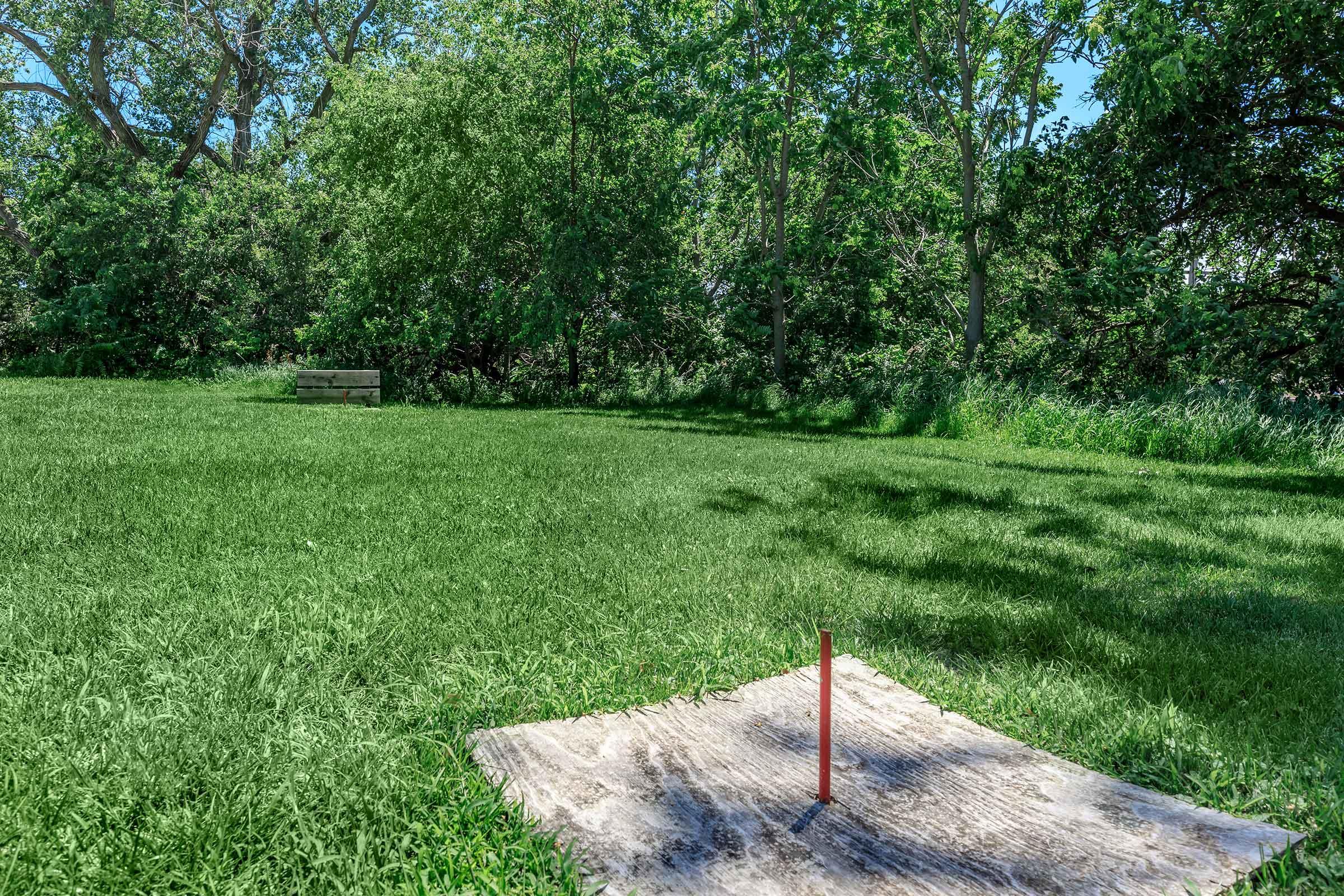 A sunny park scene featuring a patch of lush green grass, a wooden platform with a red vertical object in the foreground, and a bench in the background surrounded by trees. The overall atmosphere is calm and serene, showcasing nature and open space.