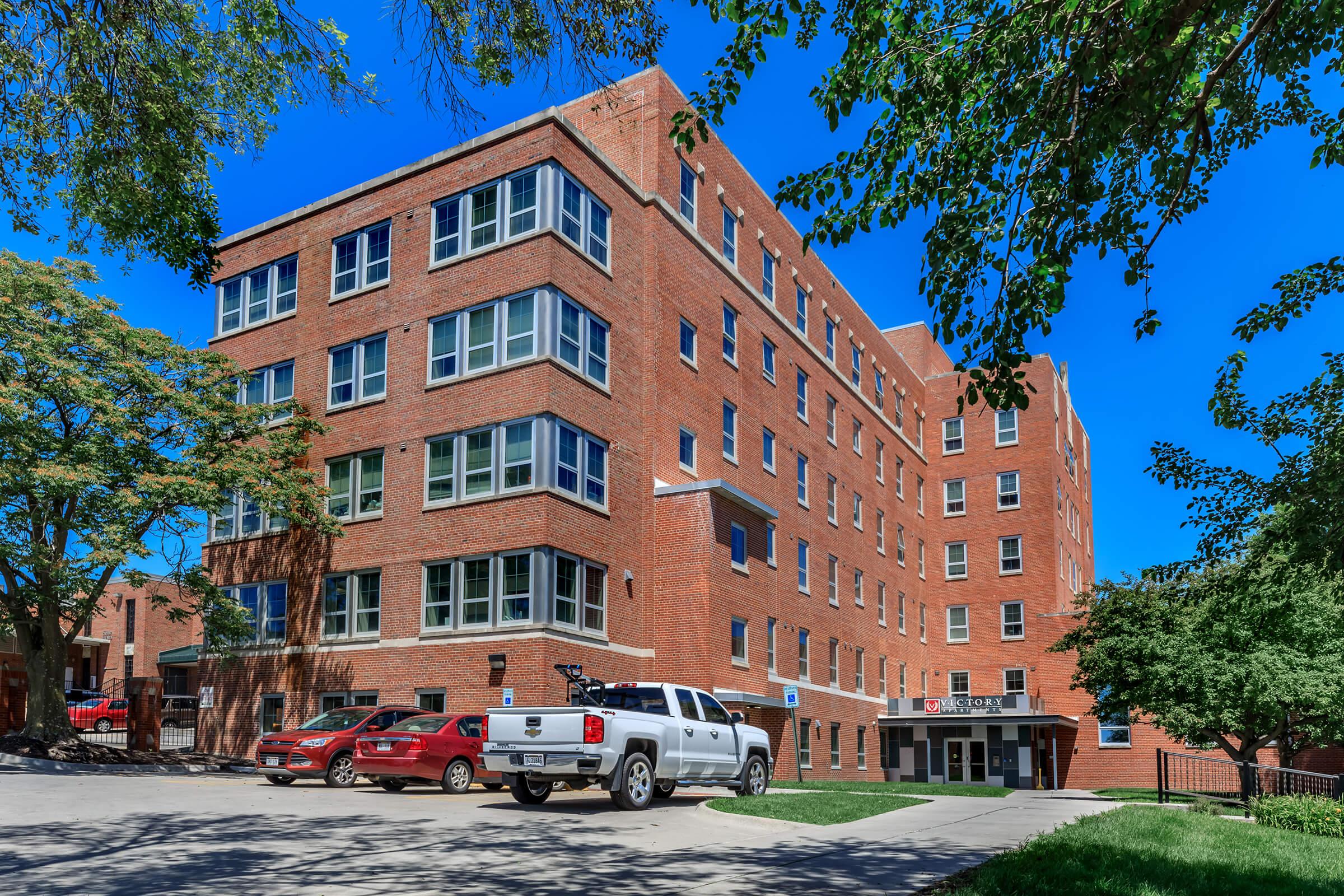 A large red brick building with multiple stories, featuring numerous windows. In front, there are several parked cars including a white truck and red vehicles. The area is surrounded by trees under a clear blue sky.