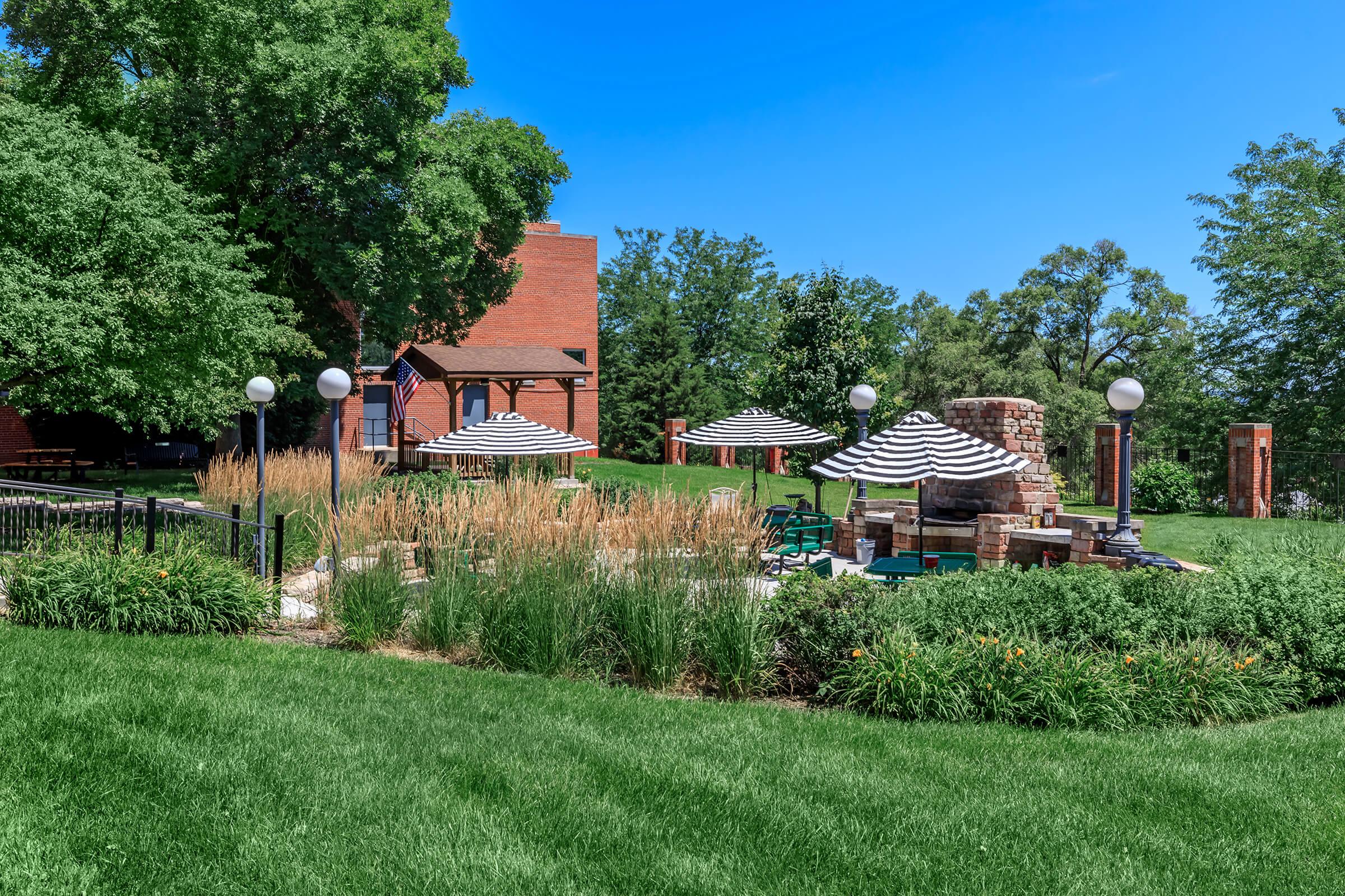 A scenic outdoor area featuring green grass, tall ornamental grass, and striped umbrellas over seating areas. There is a stone fire pit and a brick building in the background, all under a clear blue sky with some trees nearby, creating a relaxing atmosphere for gatherings.