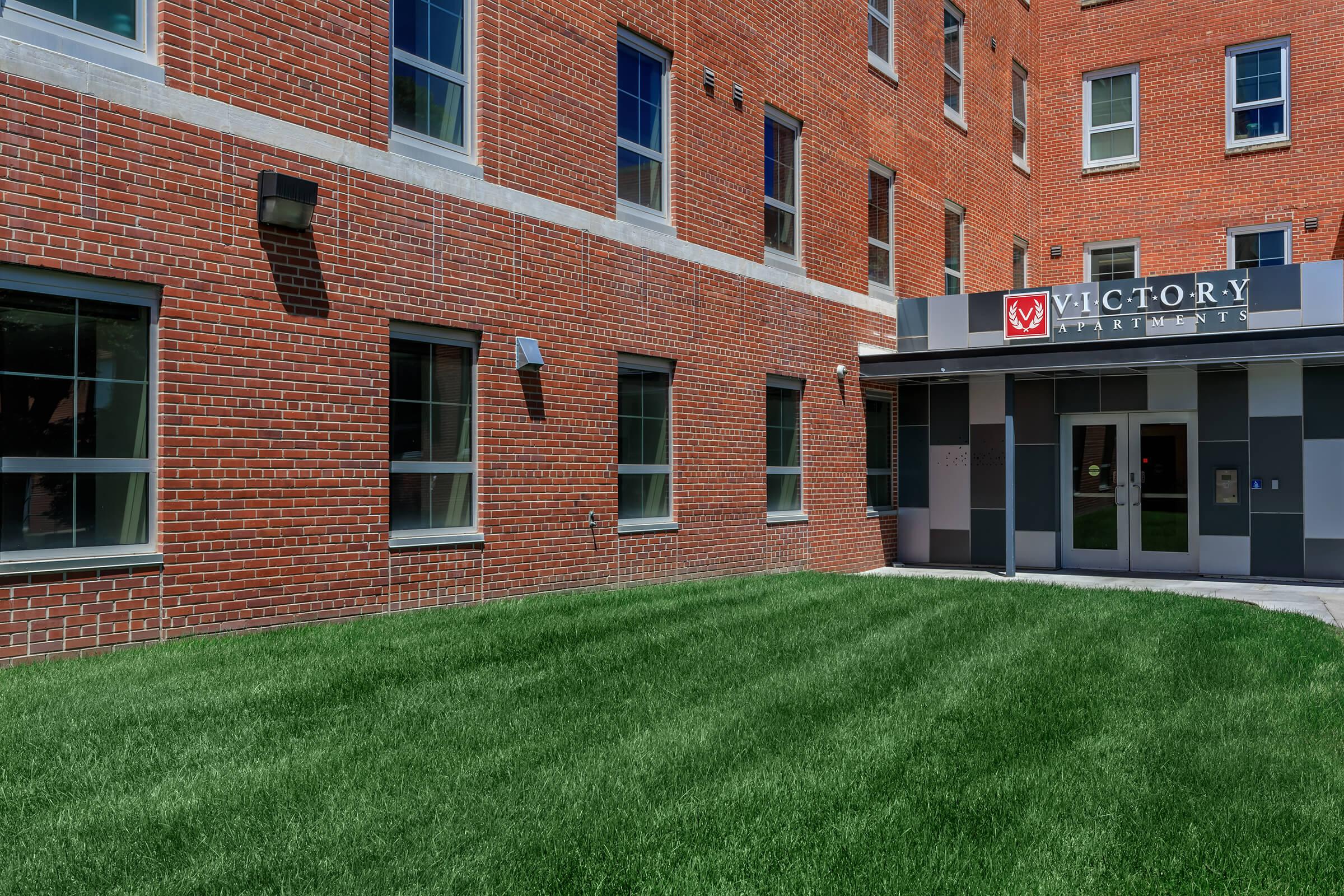 Exterior view of Victory Apartments, featuring a modern entrance with a sign. The building has a brick facade and large windows. A well-maintained green lawn is in the foreground, complementing the contemporary design of the apartment complex.