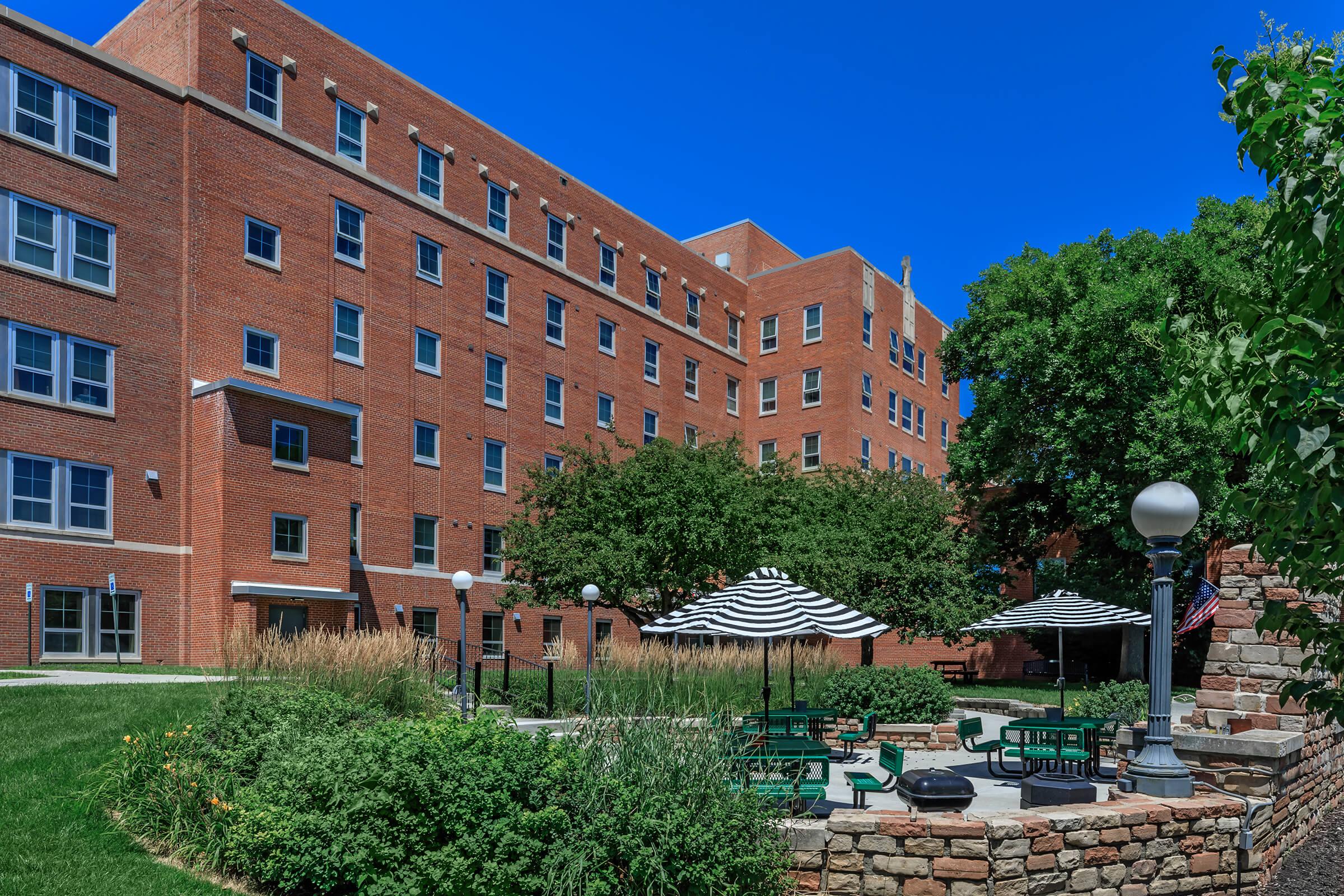 A sunny view of a brick building with multiple windows, surrounded by greenery. In the foreground, there are picnic tables under striped umbrellas, enhancing the inviting outdoor space. Lush grass and tall plants add to the pleasant atmosphere.