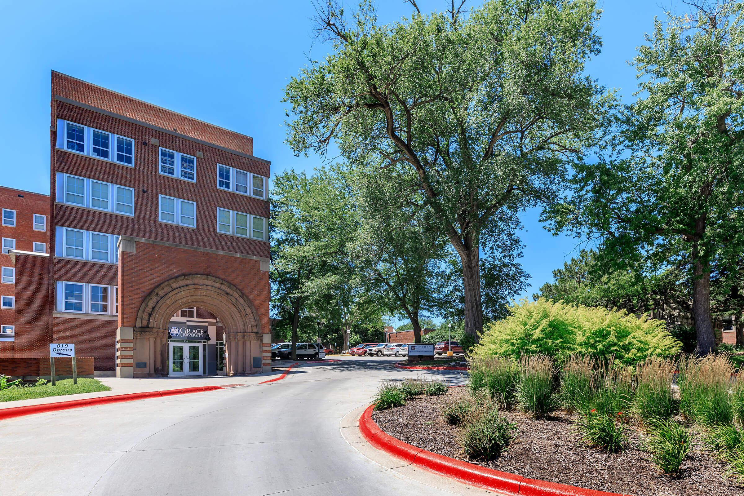 A well-maintained building with a brick façade and large windows, surrounded by lush green trees. The driveway curves gently, flanked by landscaped areas with grass and ornamental plants. A clear blue sky adds to the bright and inviting atmosphere of the scene.