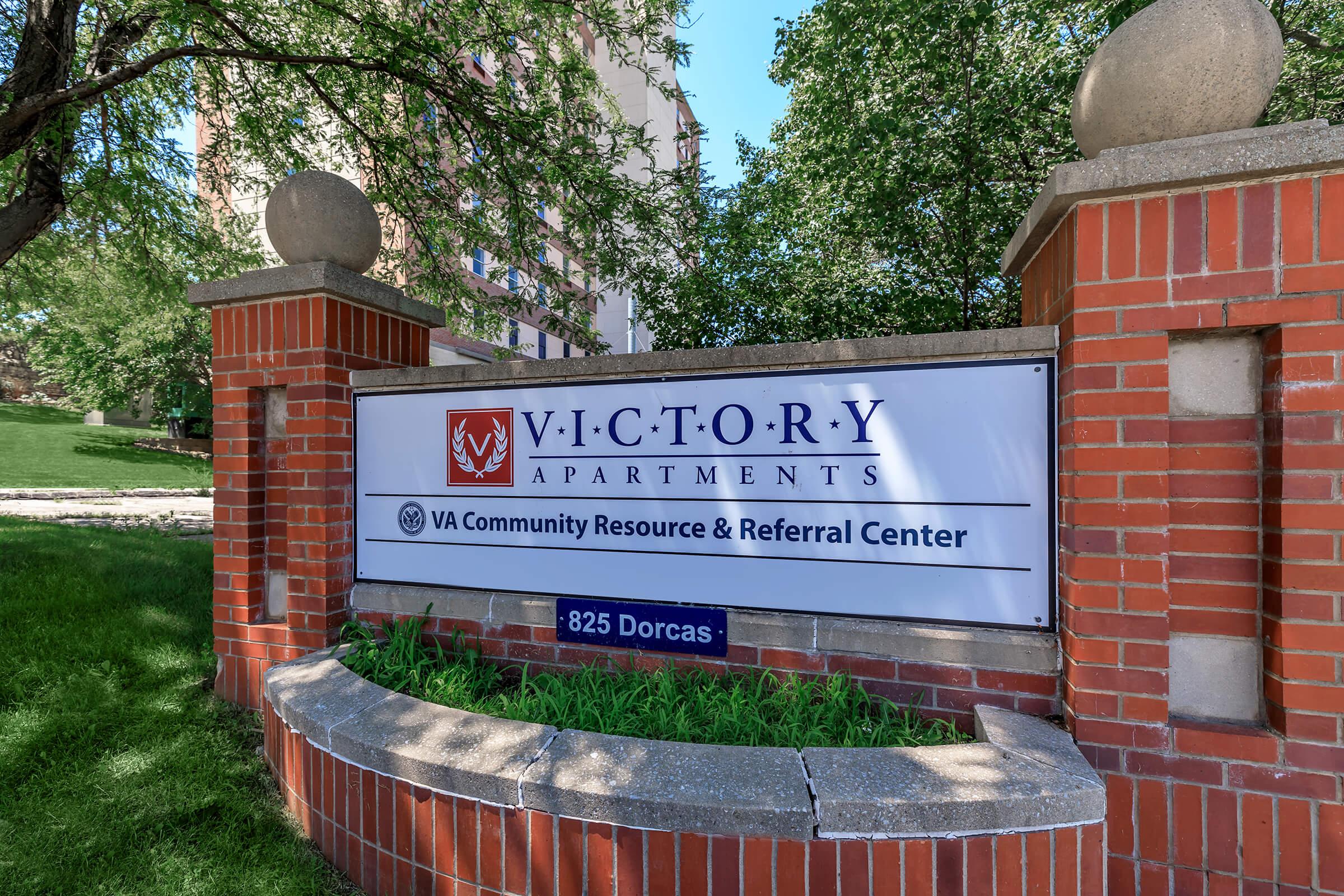 Sign for Victory Apartments featuring the VA Community Resource & Referral Center, located at 825 Dorcas. The sign is mounted on a brick structure surrounded by greenery.