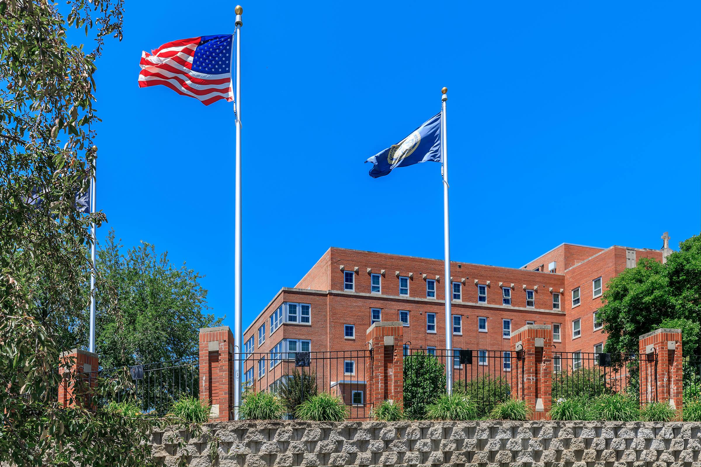 Two flagpoles stand in front of a brick building. The left flagpole features the American flag, while the right displays a state flag. The building, characterized by multiple windows, is surrounded by greenery and a stone wall, set against a clear blue sky.
