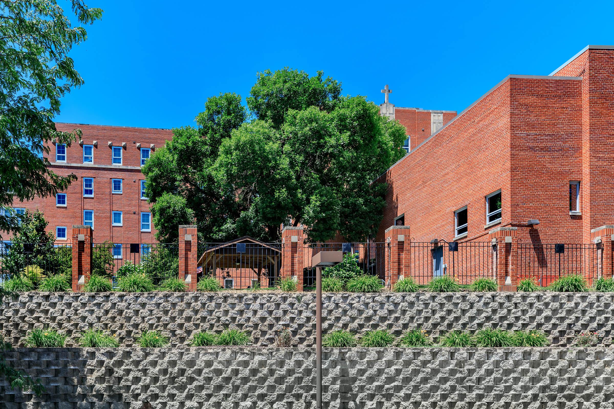 A brick building with a large tree in the foreground and a fenced area. The scene features a blue sky, greenery along a stone wall, and a visible cross atop a structure in the background.