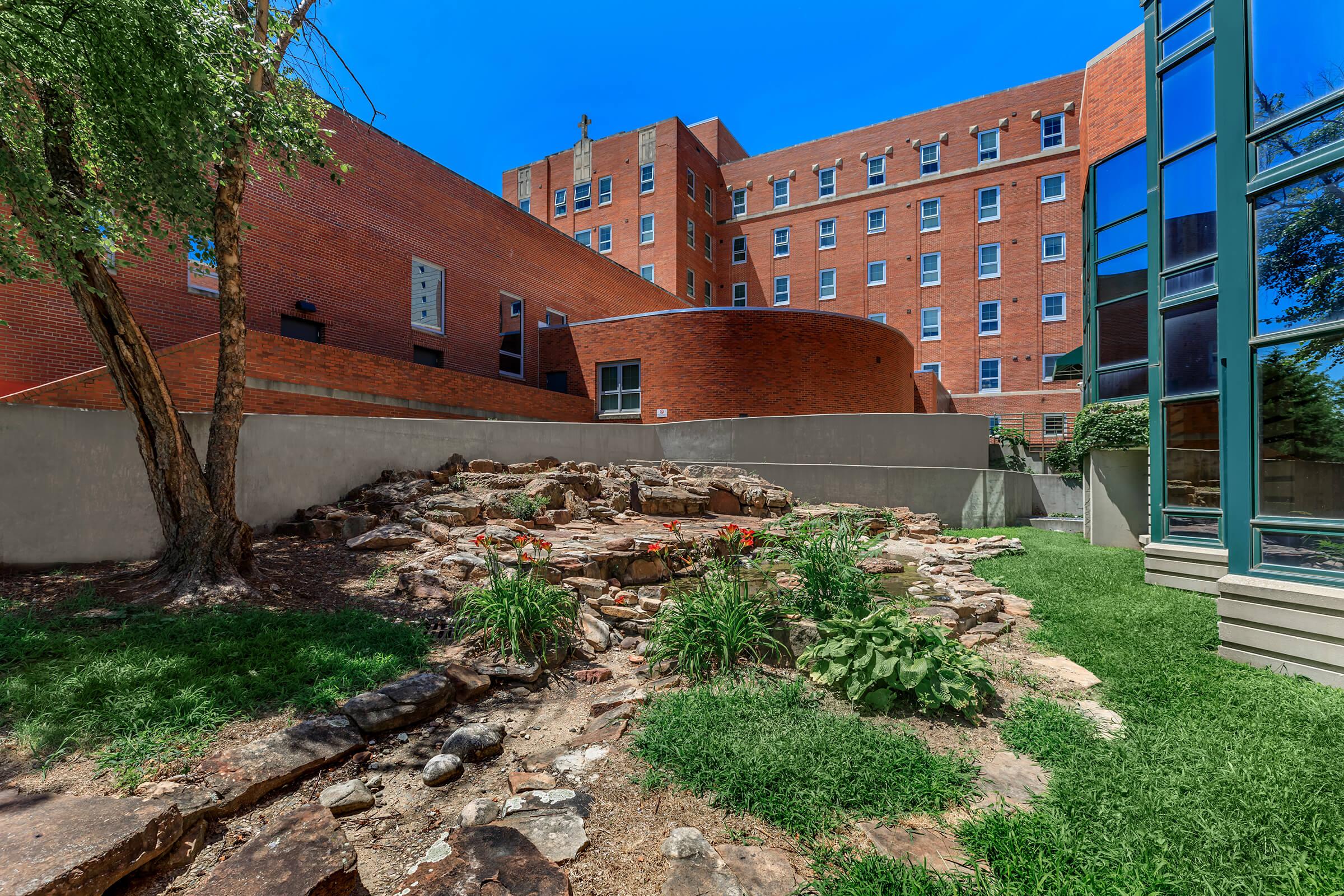 A landscaped area featuring a small rocky garden with greenery, surrounded by a stone wall. In the background, a large red-brick building and a modern glass structure are visible under a clear blue sky. The scene captures a blend of nature and architecture.
