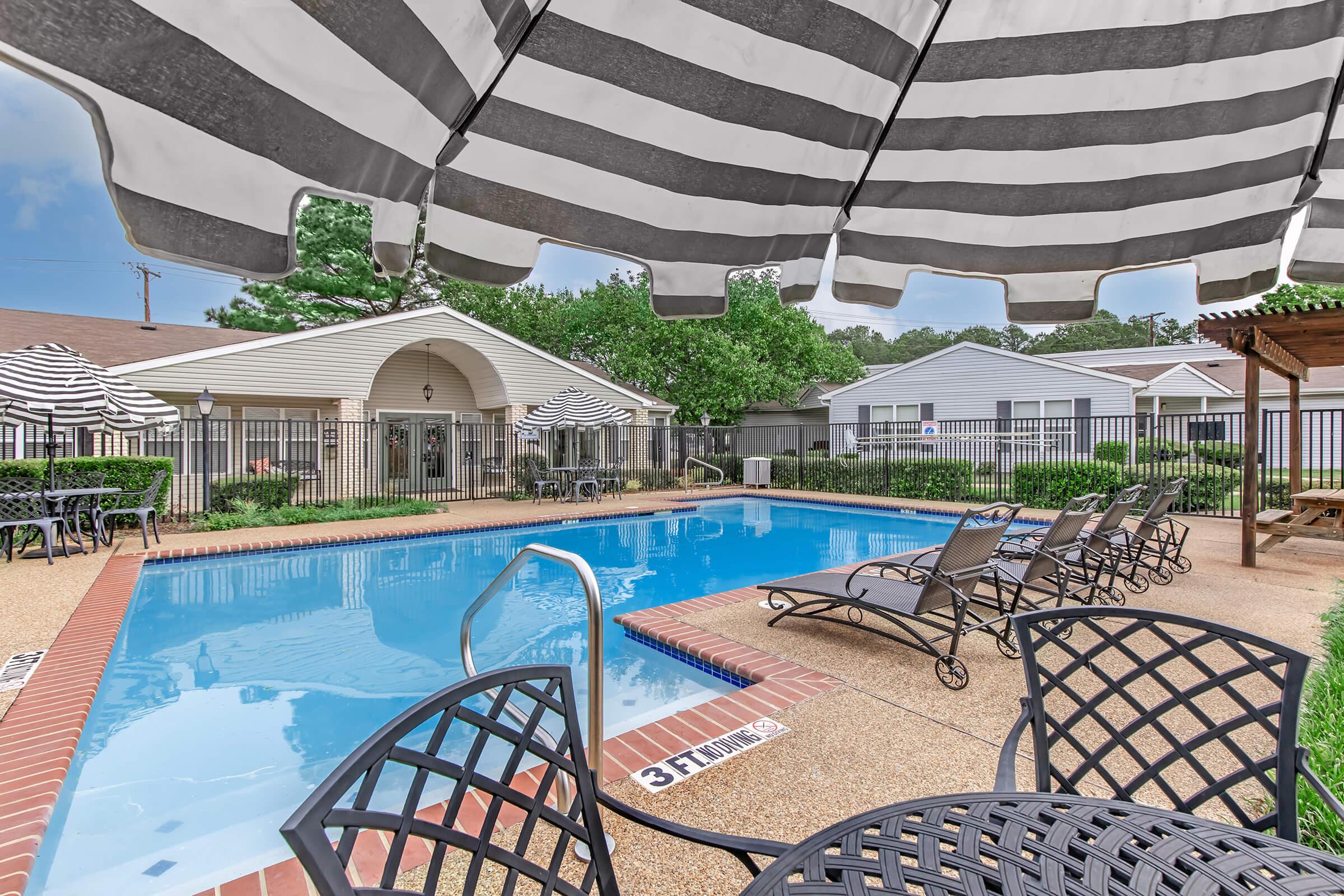 A swimming pool surrounded by lounge chairs and tables under a striped umbrella. The pool area is enclosed by a fence, with trees and buildings in the background. The setting is peaceful and inviting, perfect for relaxation and leisure.