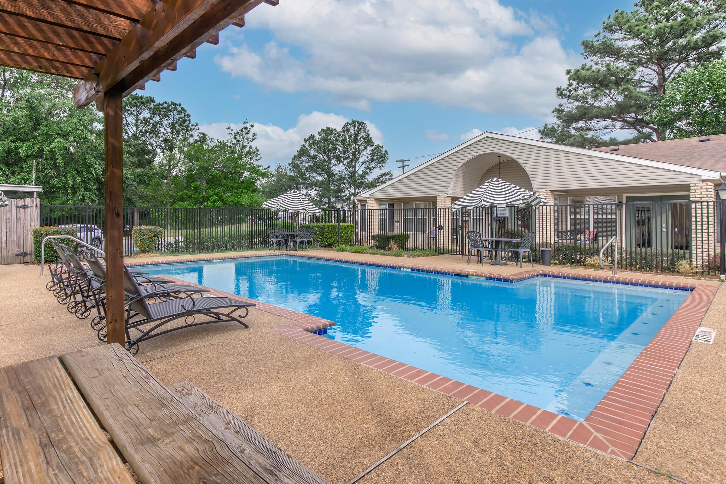 A serene pool area featuring a crystal clear blue pool surrounded by lounge chairs. Nearby, there are shaded tables with umbrellas and a well-maintained fence. The background includes greenery and a residential building with a light-colored facade, all under a partly cloudy sky.
