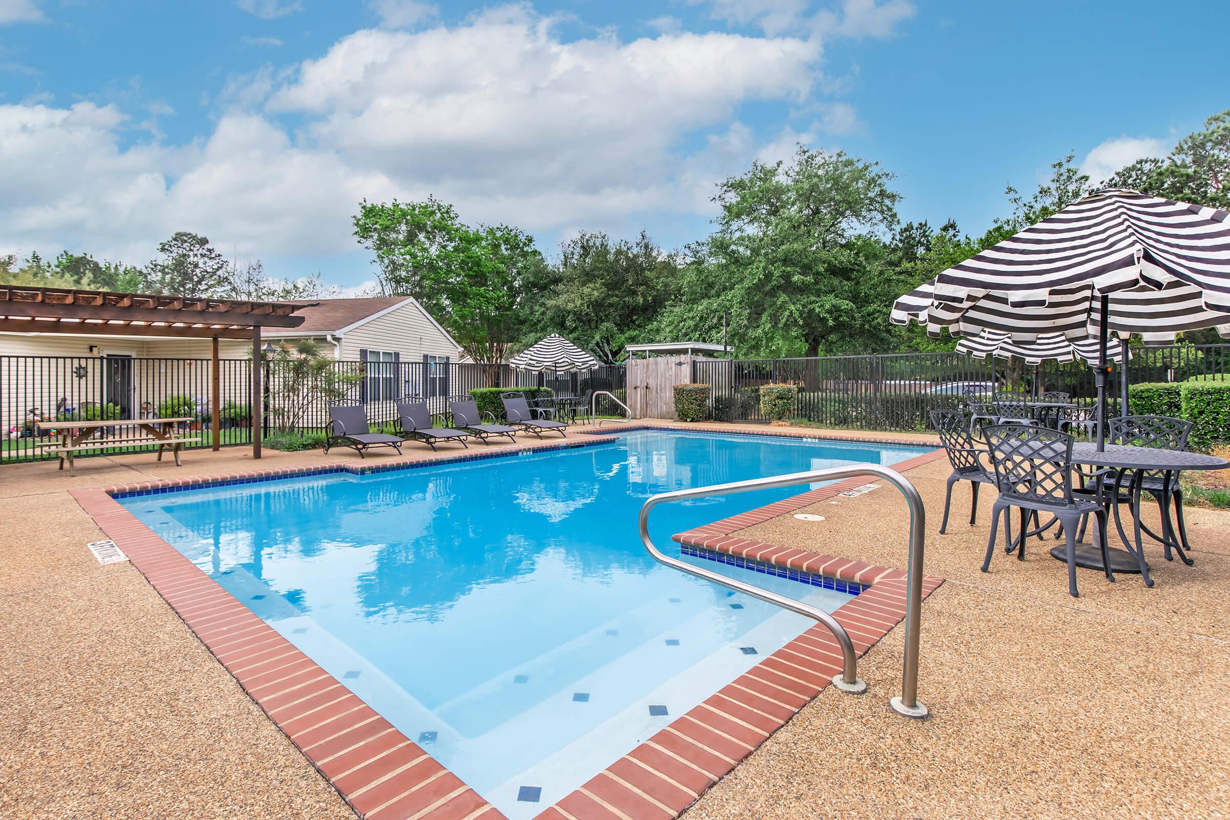 A clear blue swimming pool surrounded by lounge chairs and tables with umbrellas. Lush green trees and a fence provide a nice backdrop. The area is well-maintained, inviting for relaxation and leisure activities.