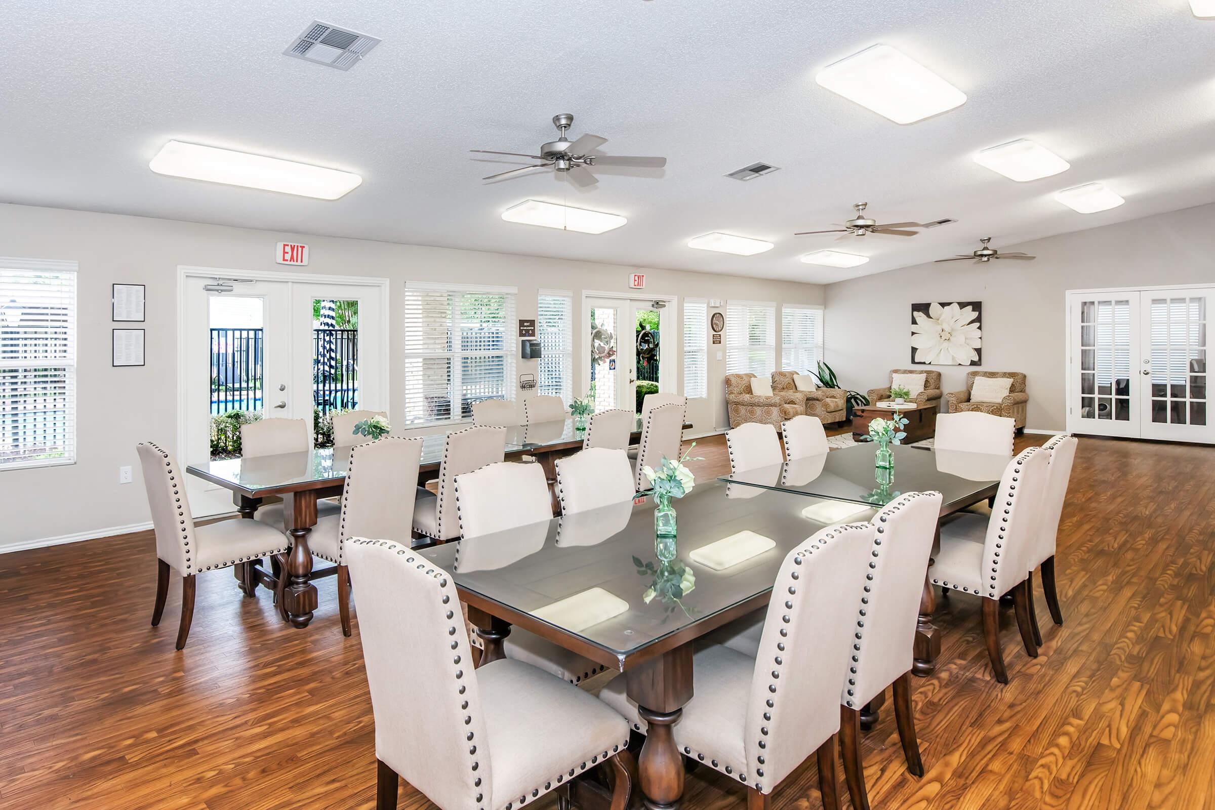 A well-lit communal dining area featuring a long table surrounded by upholstered chairs. The space has light-colored walls, ceiling fans, and large windows that let in natural light. Potted plants add a touch of greenery, and there are comfortable seating areas with couches in the background.