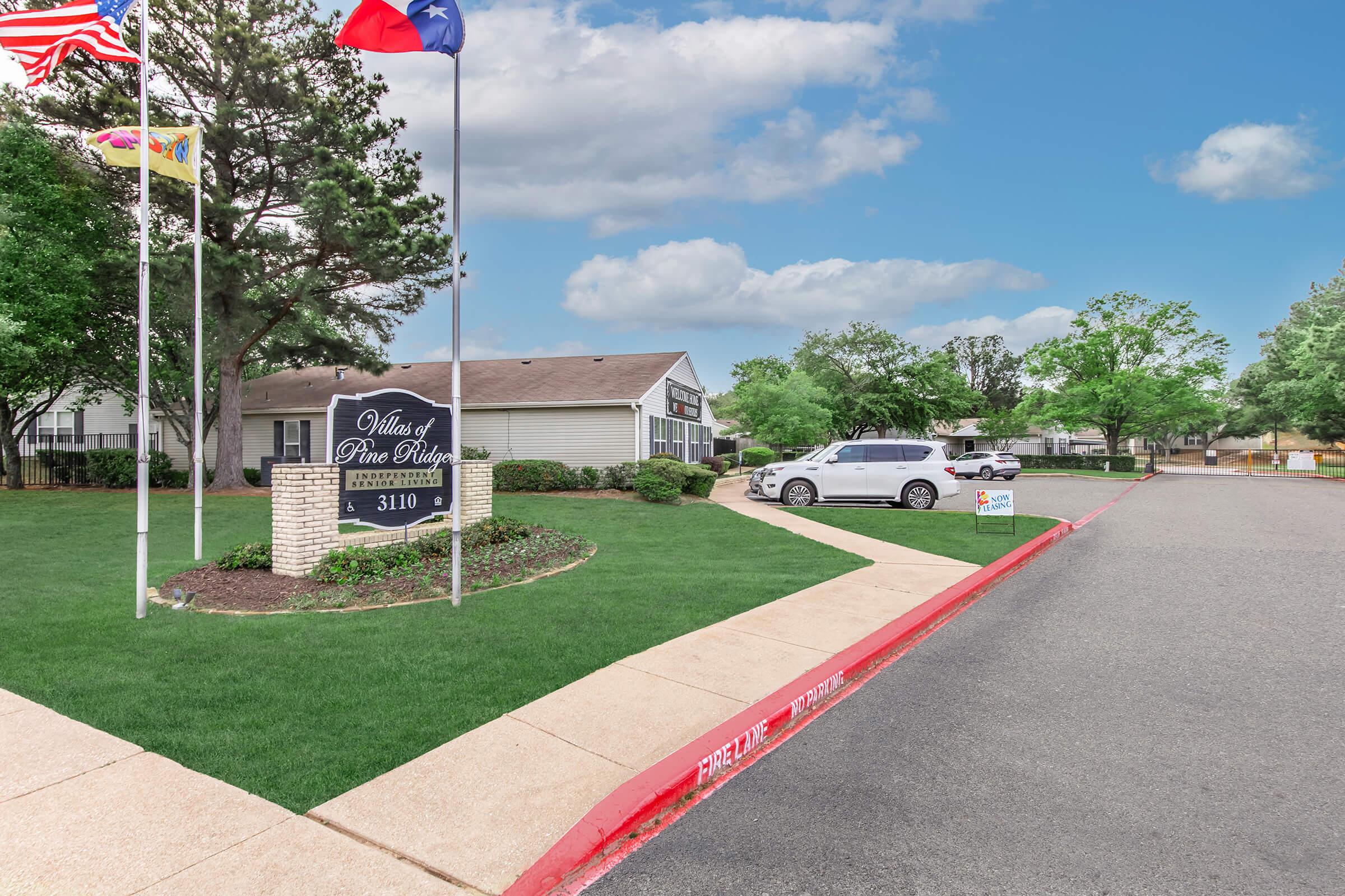 A view of the entrance to "Mills of Pine Ridge," featuring a signpost with flags. The scene includes an inviting landscape with well-maintained grass, trees, and a clear sky. A white vehicle is parked nearby, and there is a parking area with a red curb marking. The setting conveys a welcoming atmosphere for residents and visitors.
