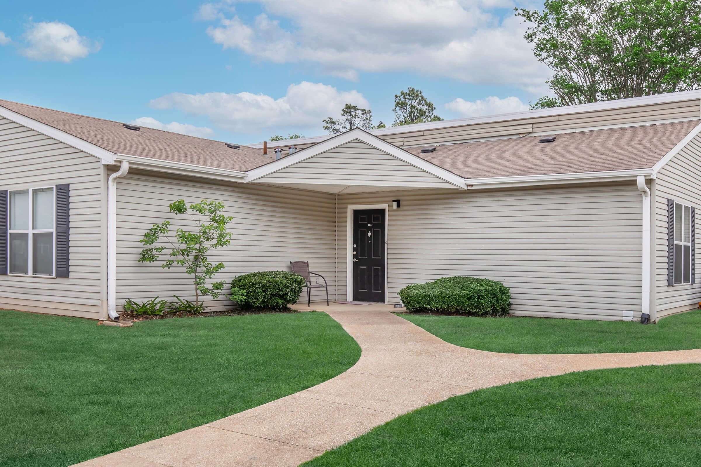 A well-maintained single-story home with a light-colored exterior and a neatly landscaped front yard. The path leading to the front door is bordered by green grass and a small bush. The sky is clear with a few clouds, enhancing the inviting atmosphere of the property.