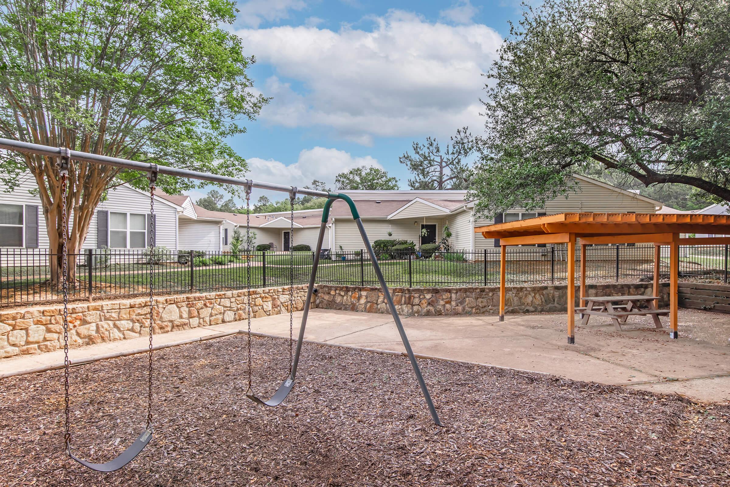A playground area featuring a swing set with two empty swings, a wooden picnic table under a gazebo-like structure, and a landscaped yard with trees and small bushes. The background includes several residential buildings and a clear sky with scattered clouds.