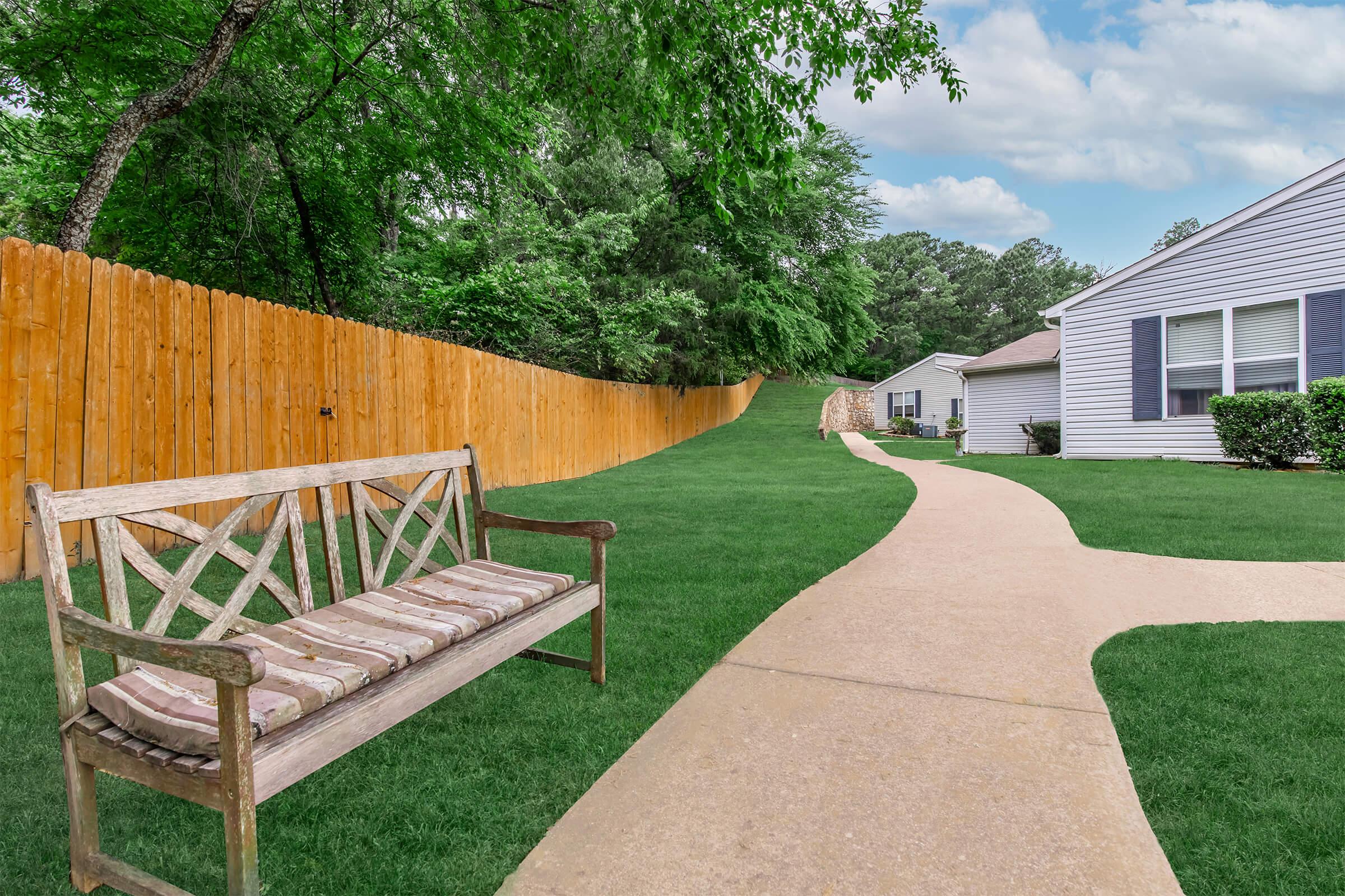 A view of a landscaped area featuring a wooden bench on a grassy lawn, bordered by a wooden fence. A paved pathway winds through the space, leading towards a residential building with a garden area. The scene is serene, surrounded by trees under a partly cloudy sky.