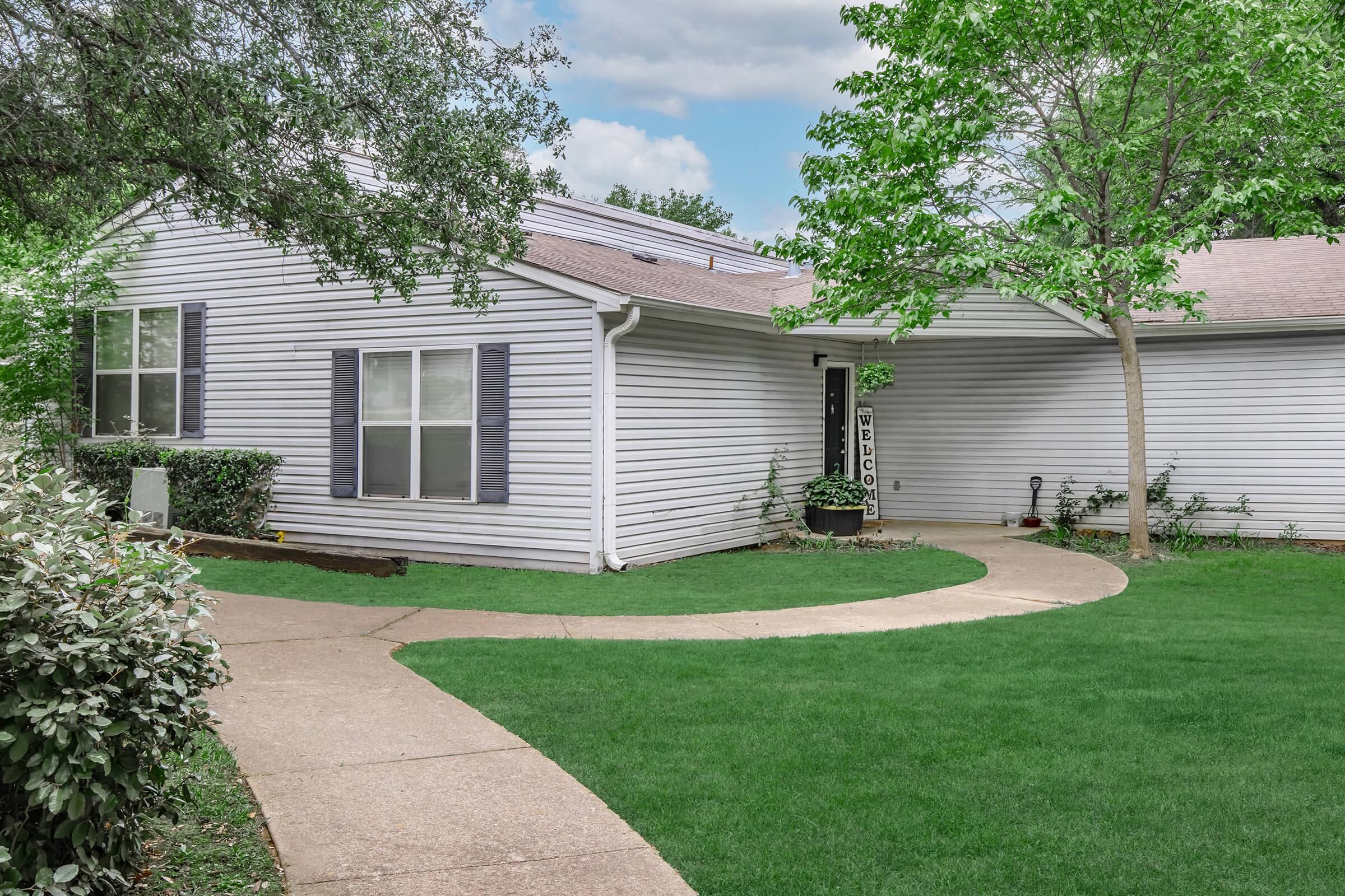A light-colored single-story house with blue shutters, surrounded by lush greenery. A curved walkway leads to the front door, which is adorned with a welcome sign. Trees and well-maintained shrubs frame the entrance, creating a welcoming atmosphere in a residential area.