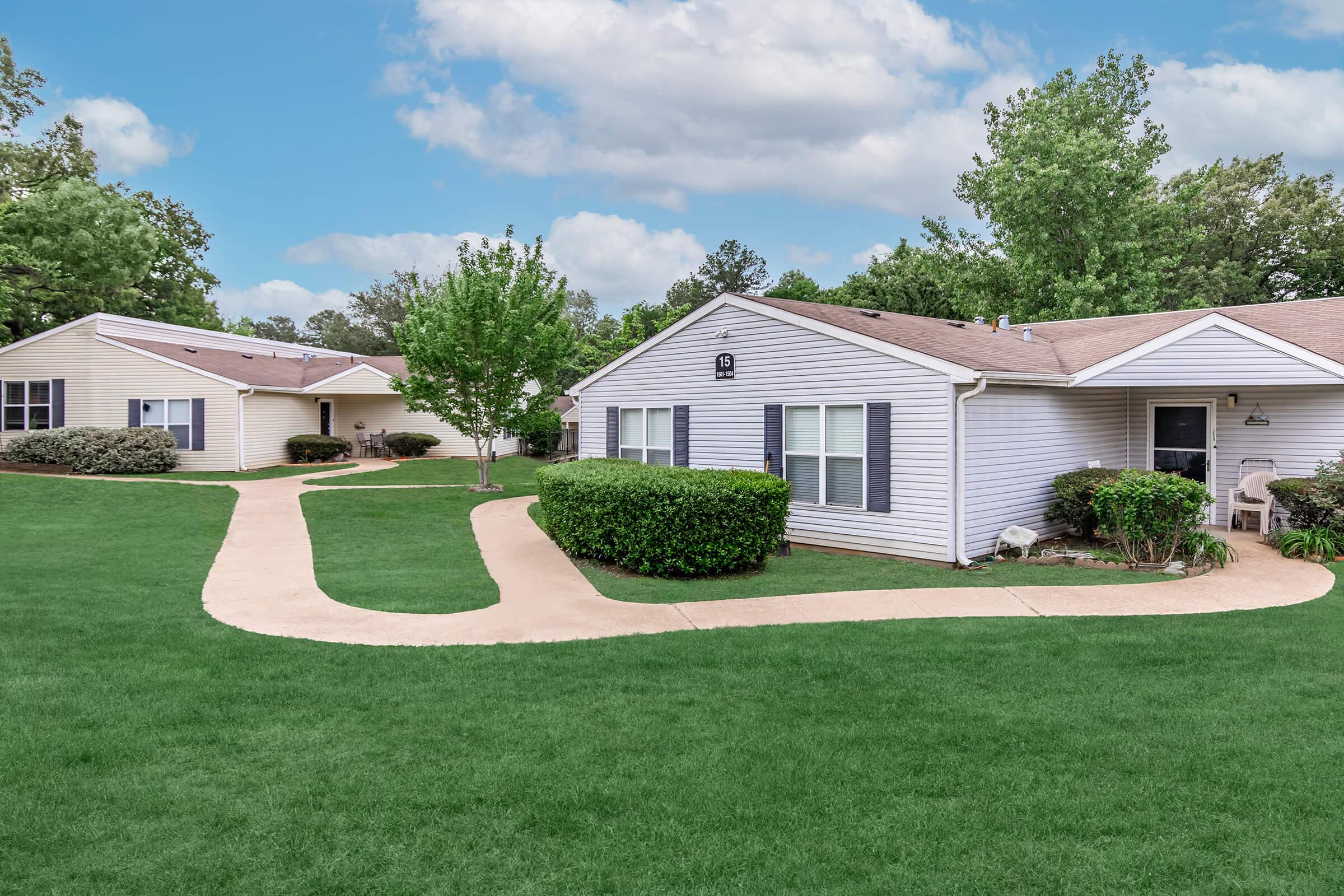 A scenic view of two single-story residential buildings surrounded by well-maintained lawns and trees. A curved pathway connects the buildings, providing a serene, inviting atmosphere on a clear day with a blue sky and fluffy clouds.