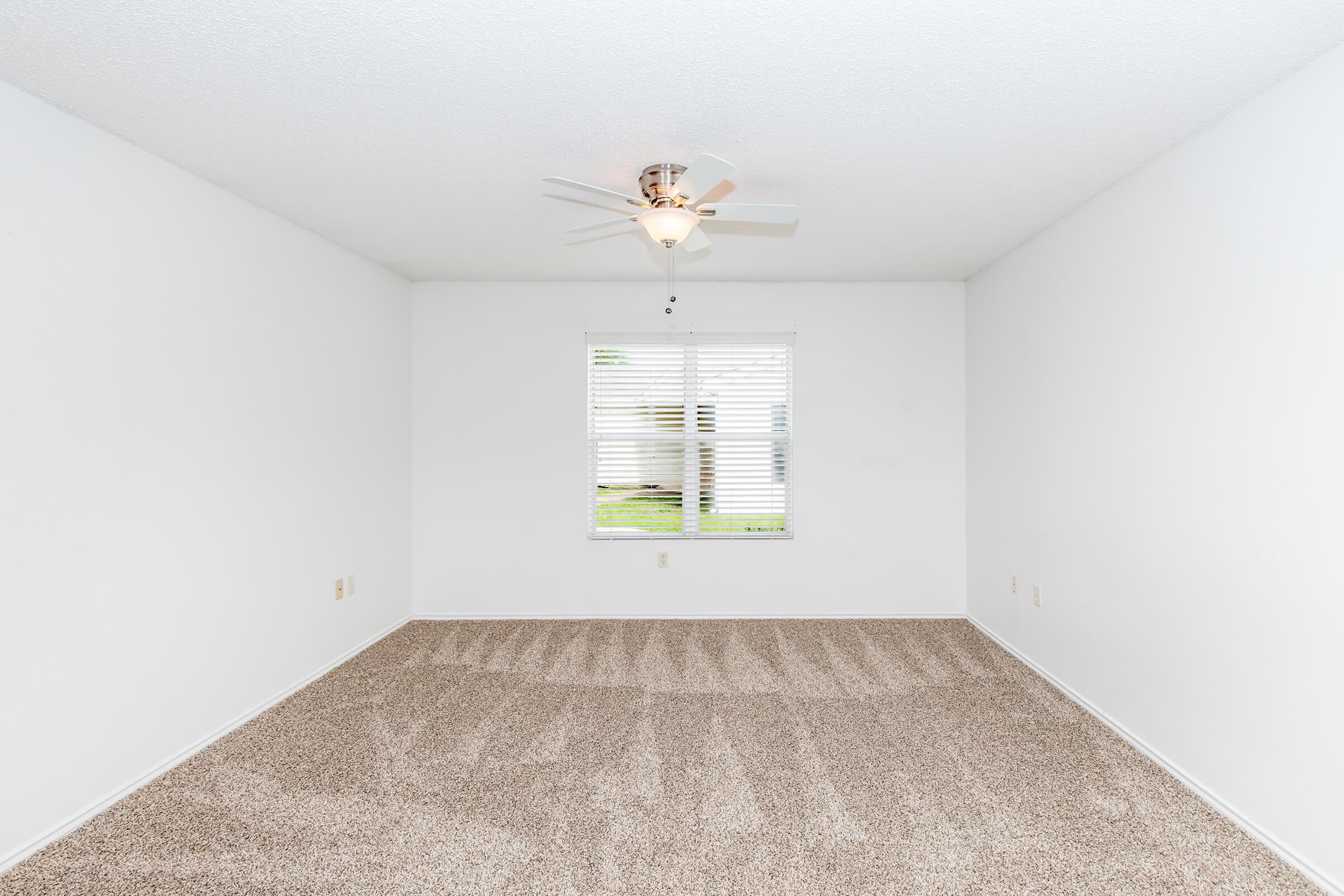 Empty room with beige carpet, a ceiling fan, and a window with blinds. The walls are white, and there are no furnishings or decorations. Natural light filters through the window, illuminating the space.