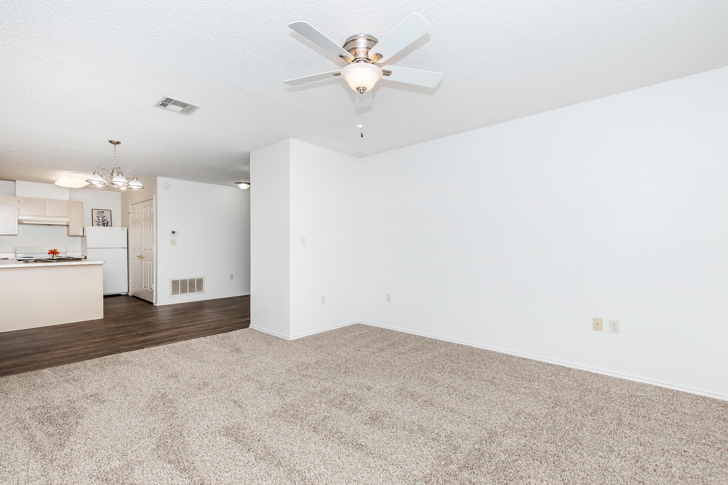A spacious, empty living room with light-colored carpet and a ceiling fan. A kitchen area is visible in the background with beige cabinets and dining lighting. The walls are painted white, creating a bright and airy atmosphere.