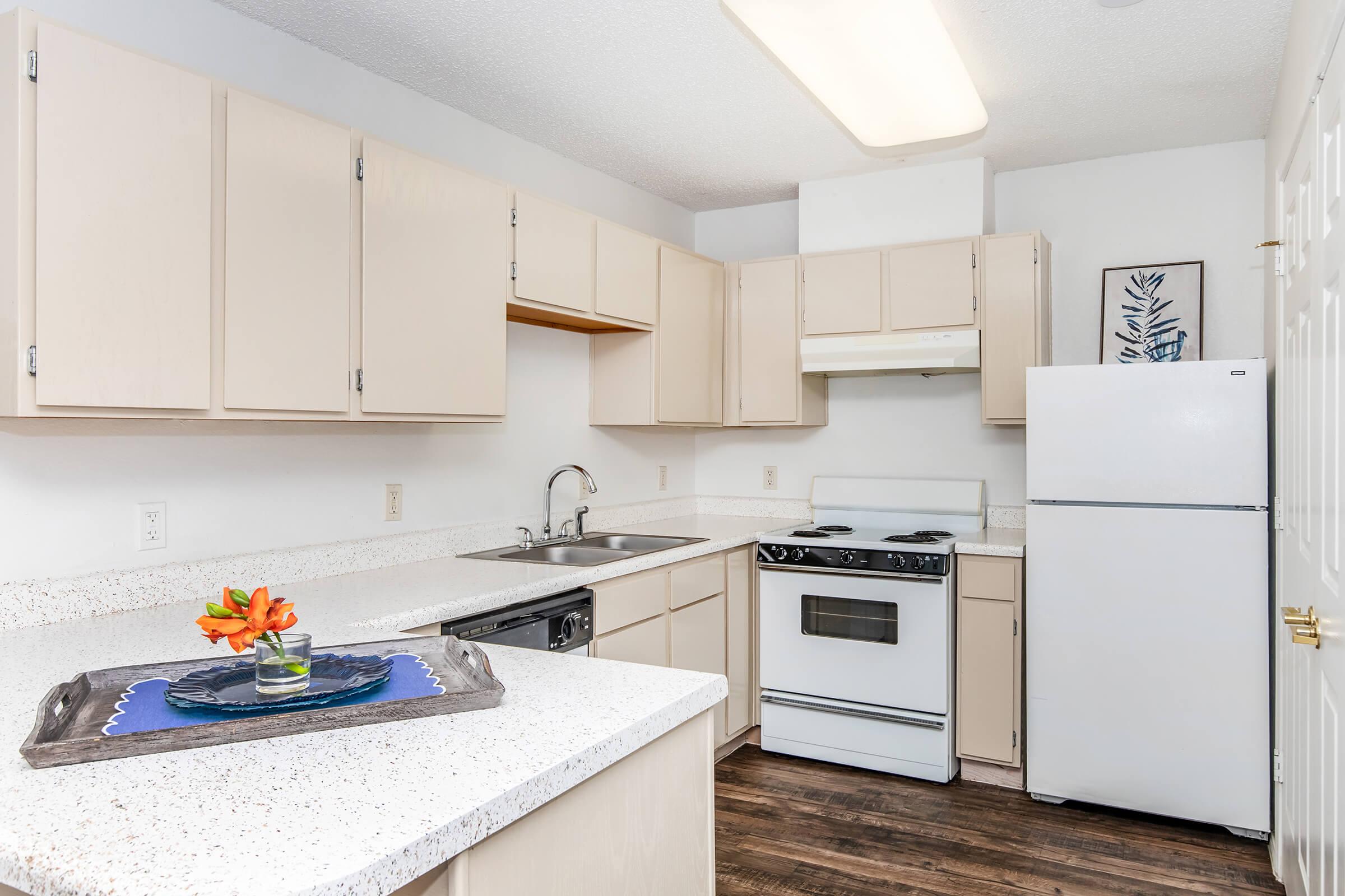 A modern kitchen with light-colored cabinets and a white refrigerator. It features a white stove, a sink, and a countertop with a tray holding a vase of orange flowers. The flooring is dark wood, and there is a framed plant illustration on the wall.
