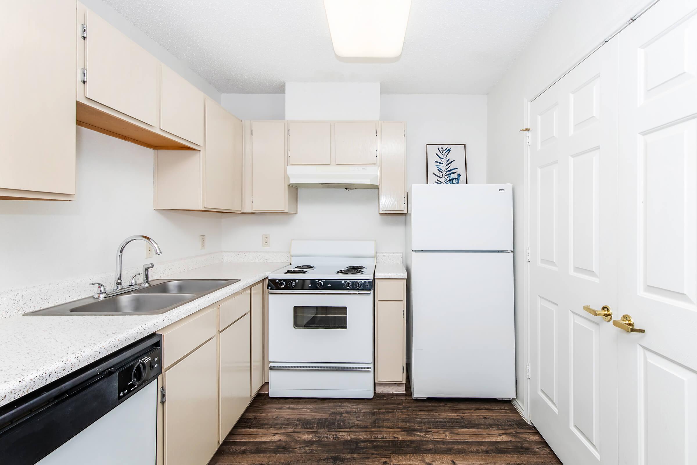 A modern kitchen featuring beige cabinets, a white refrigerator, a gas stove with oven, and a double sink. The countertops are a light-speckled finish, and the floor is dark wood. The walls are painted white, and there is a framed piece of wall art near the stove.