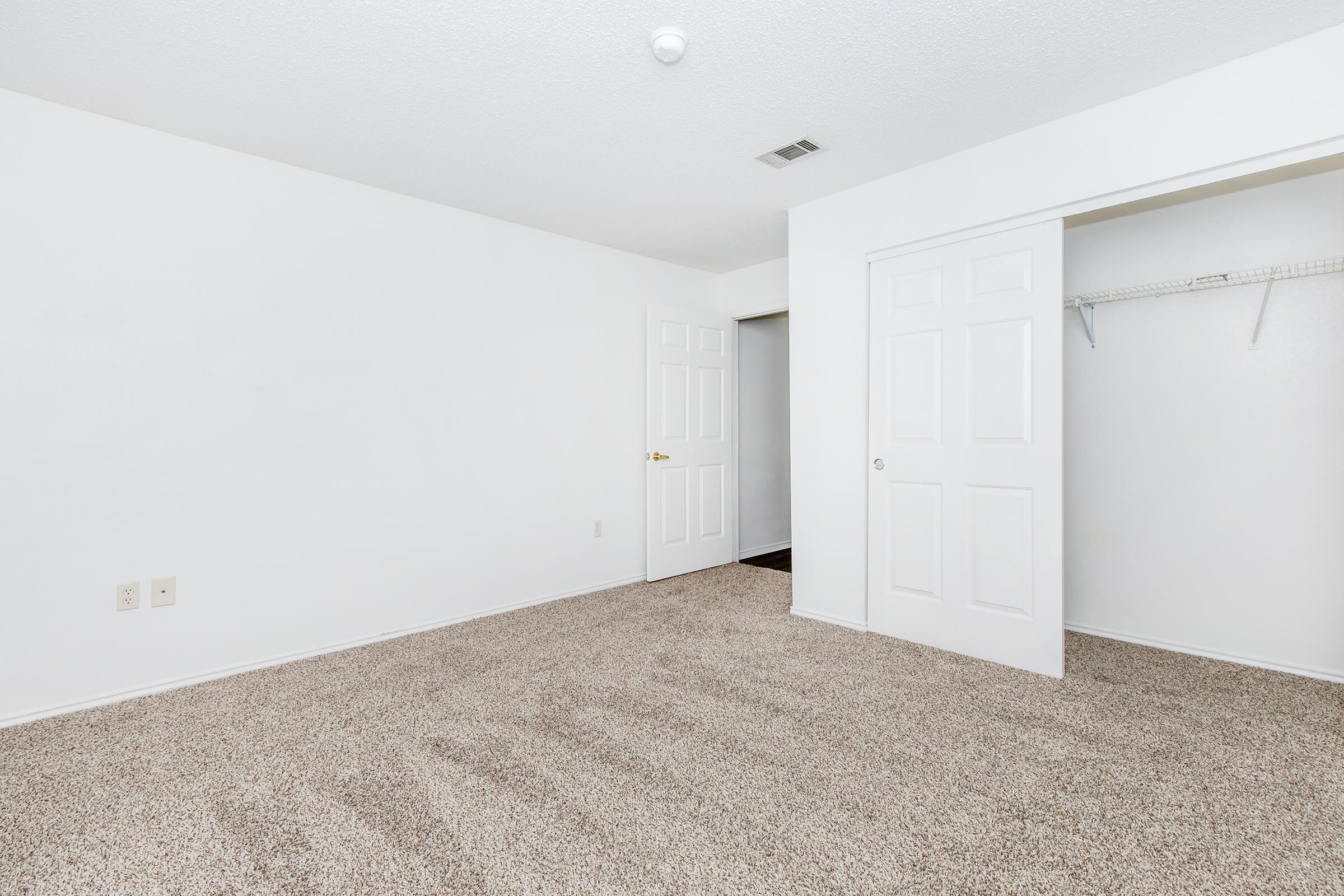 An empty room with beige carpet and white walls. A closed door on the left leads to a closet, which has a shelf and hanging space. The room is well-lit with a ceiling light, and there is a door on the right leading to another space. The overall look is minimalist and clean.