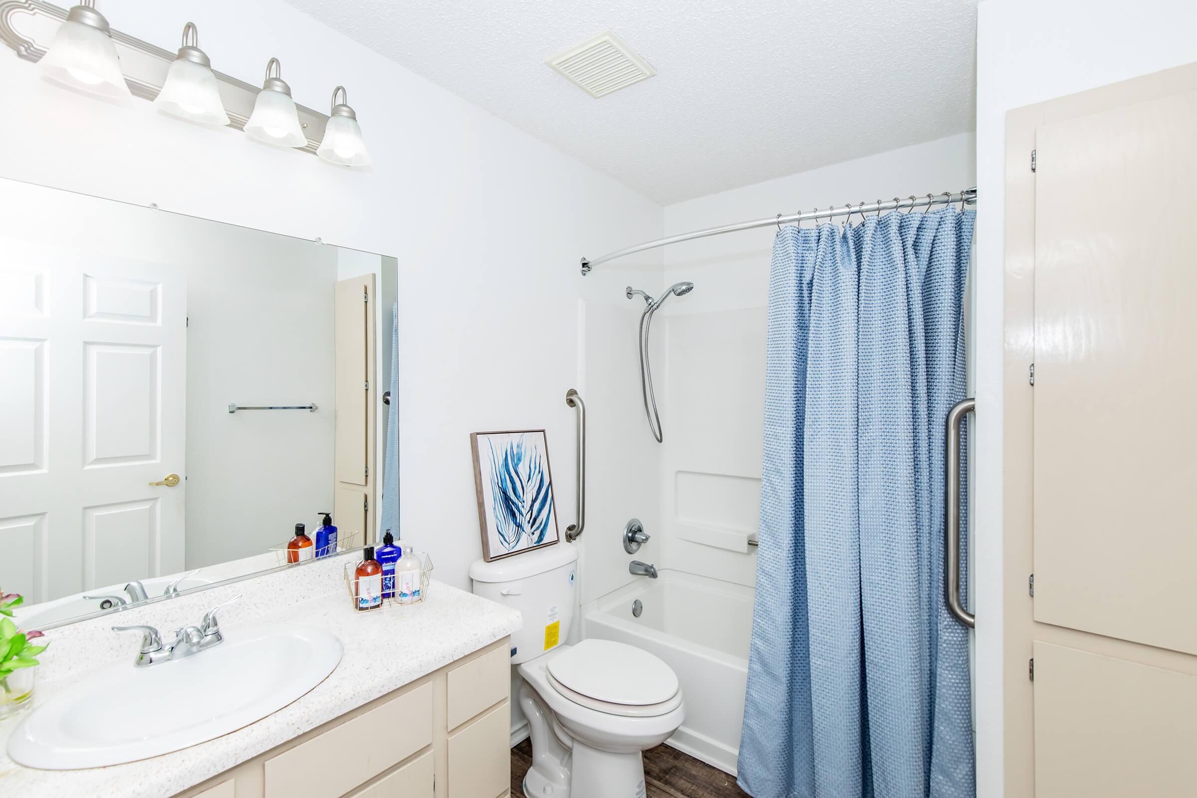A clean and modern bathroom featuring a white sink with a countertop, a mirror above it, and a shower area with a blue curtain. There is a toilet next to the shower, and some decorative items on the countertop. The walls are painted white, and there is ample natural light in the space.
