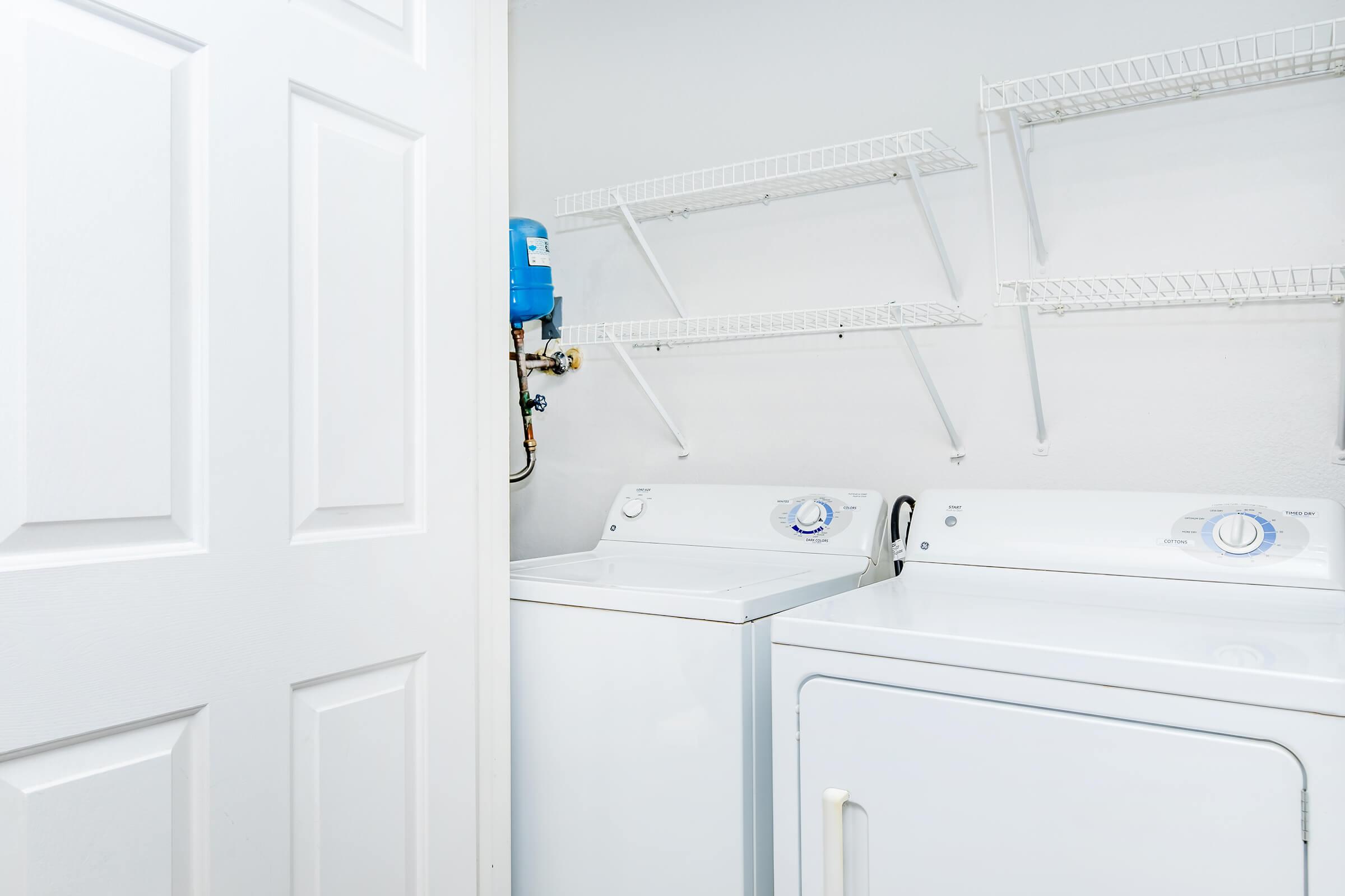 A laundry room featuring a white washing machine and dryer side by side. Above them are wire shelves mounted on the wall, providing storage space. The room has light-colored walls and a closed white door partially in view. The overall setting is clean and organized.