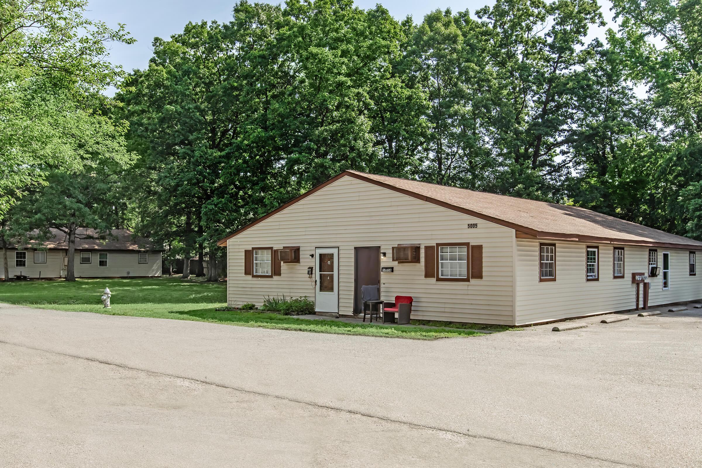 A single-story building with a light-colored exterior and brown shutters, situated in a green, tree-filled area. The structure has multiple windows and a welcoming entrance. In front, there's a red chair and a fire hydrant, with a dirt road leading to the building.