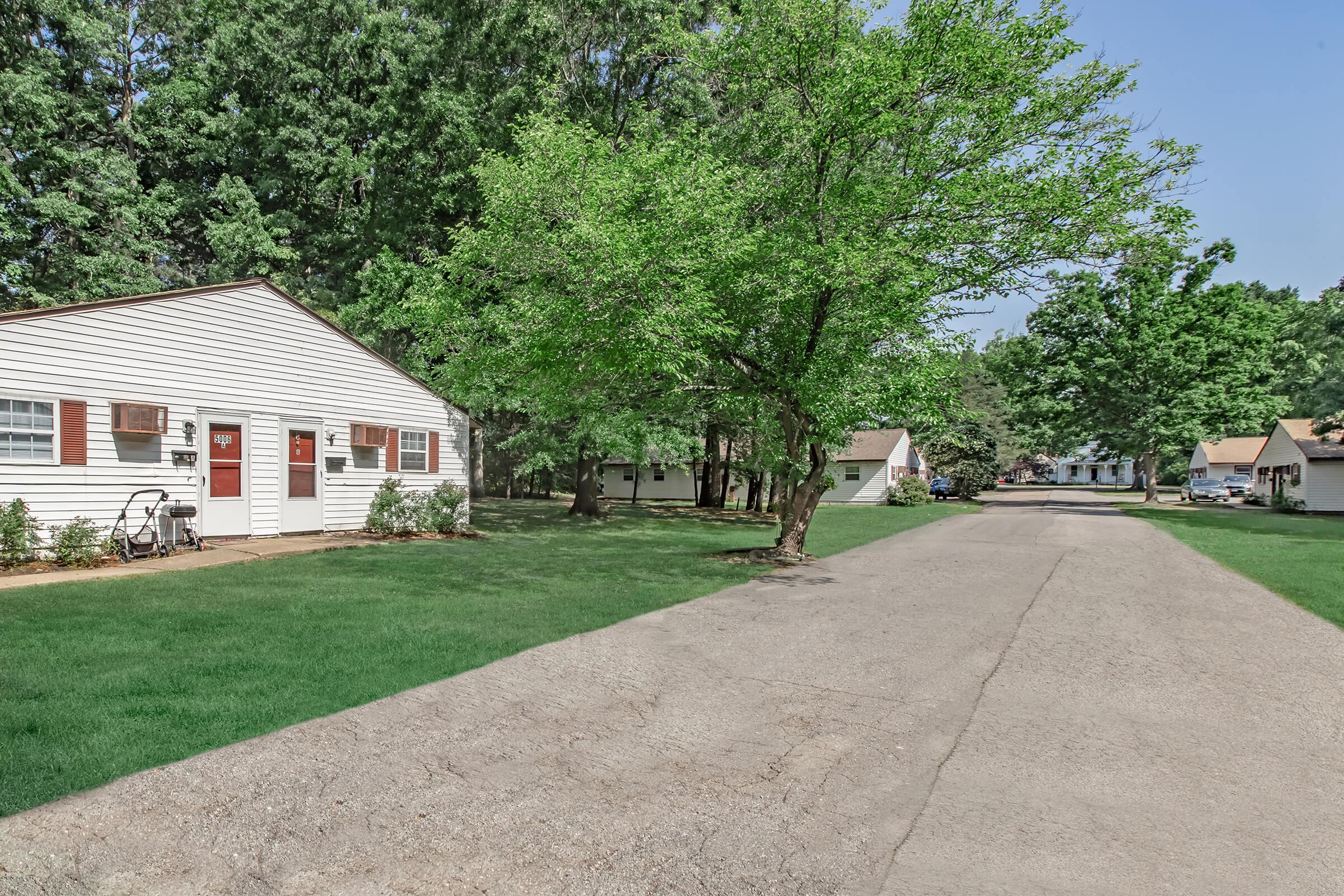 A peaceful neighborhood scene featuring several modest houses along a tree-lined street. The foreground shows a well-maintained grassy area and a paved road, while lush green trees provide shade. The houses have white siding and red doors, creating a quaint, inviting atmosphere.