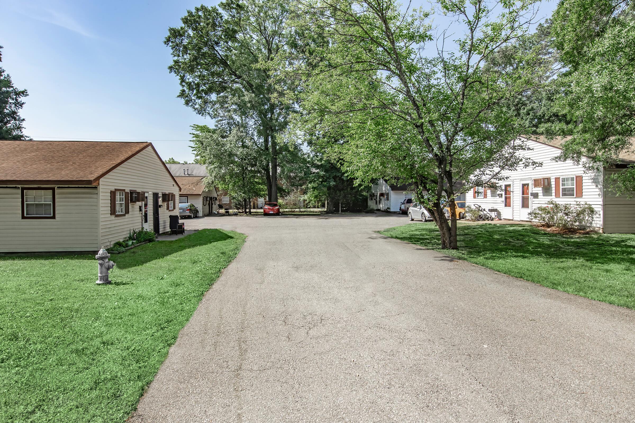 A quiet residential area showing a curved road bordered by houses and trees. On the left, there are two small homes with a fire hydrant in front. The right side features more houses, all surrounded by green grass and trees, under a clear blue sky.