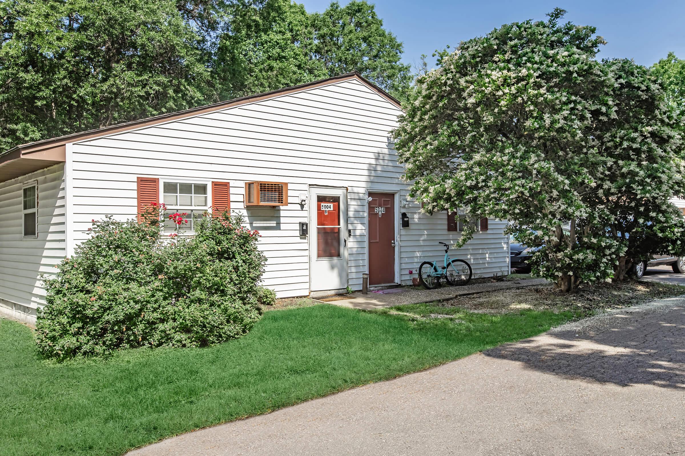 A quaint single-story house with white siding and red shutters. The front yard has green grass and a flowering shrub. There are two doors, and a bicycle is parked nearby. The setting is peaceful, surrounded by trees and a gravel path leading up to the house.