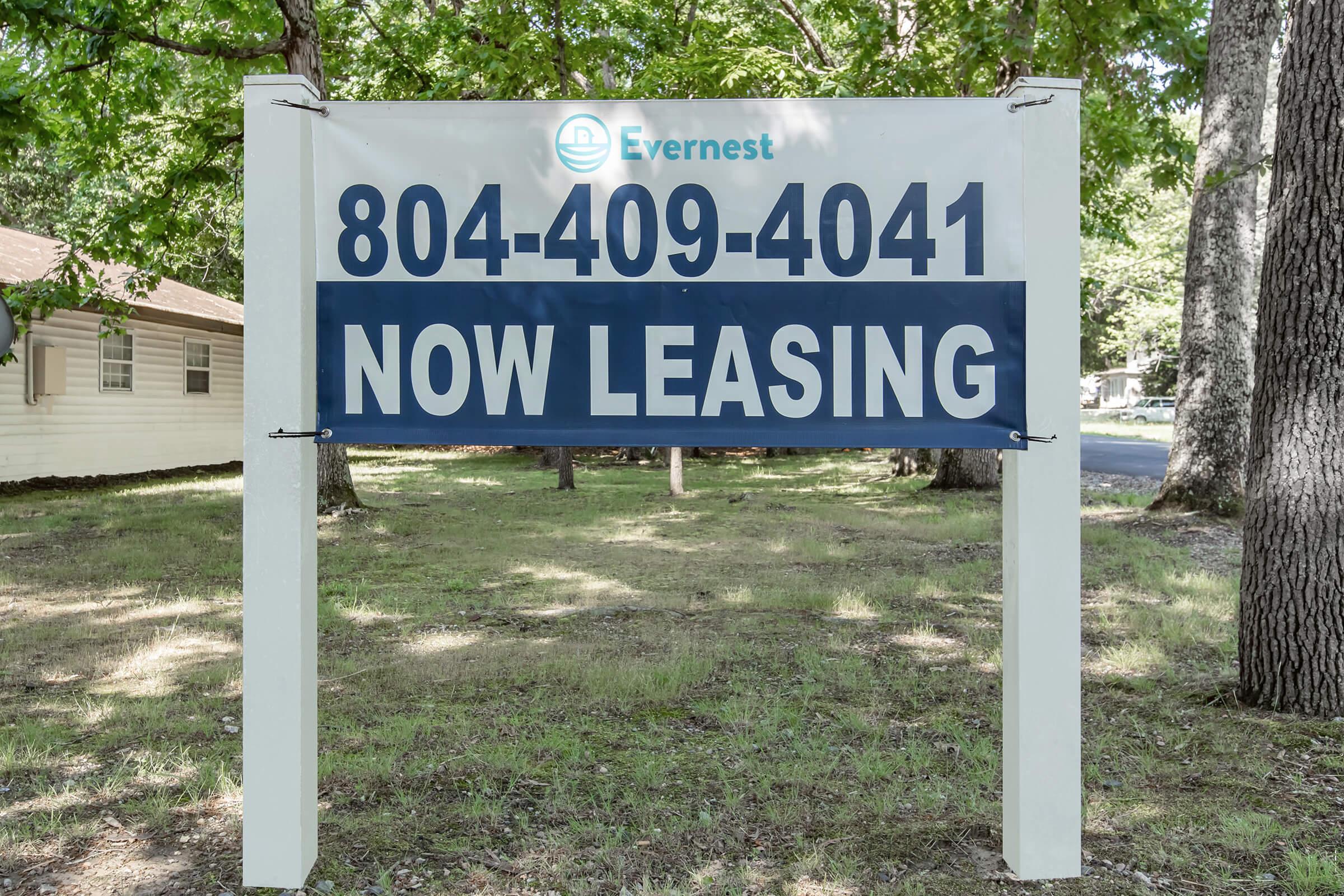 Sign indicating rental availability with the text "804-409-4041 NOW LEASING" on a blue background. The sign is surrounded by green grass and trees, suggesting a residential area.