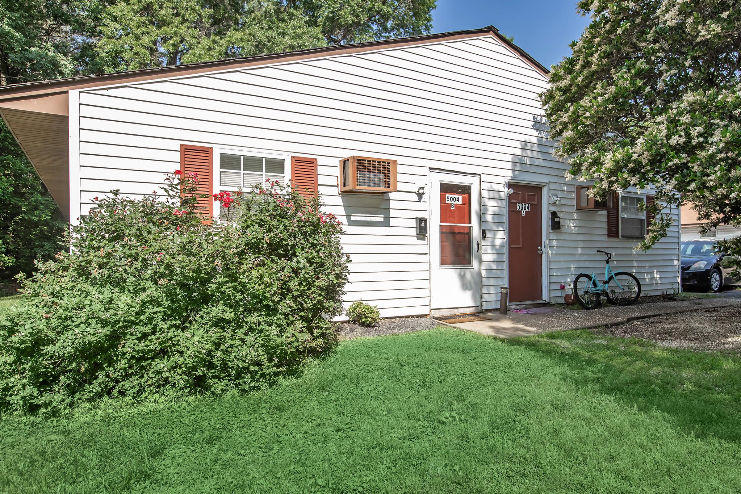 A small, single-story house with white siding and brown shutters. An air conditioning unit is visible in one of the windows. The entrance features a door with a small window, and there is a green lawn in front, along with a bush and a bicycle parked nearby.