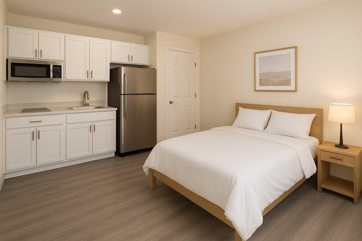 A cozy hotel room featuring a queen-sized bed with white linens, a nightstand with a lamp, a kitchenette with a microwave and refrigerator, and cabinets. The walls are painted a light color, and there is a simple framed artwork above the bed. The flooring is a light wood finish, creating a warm atmosphere.