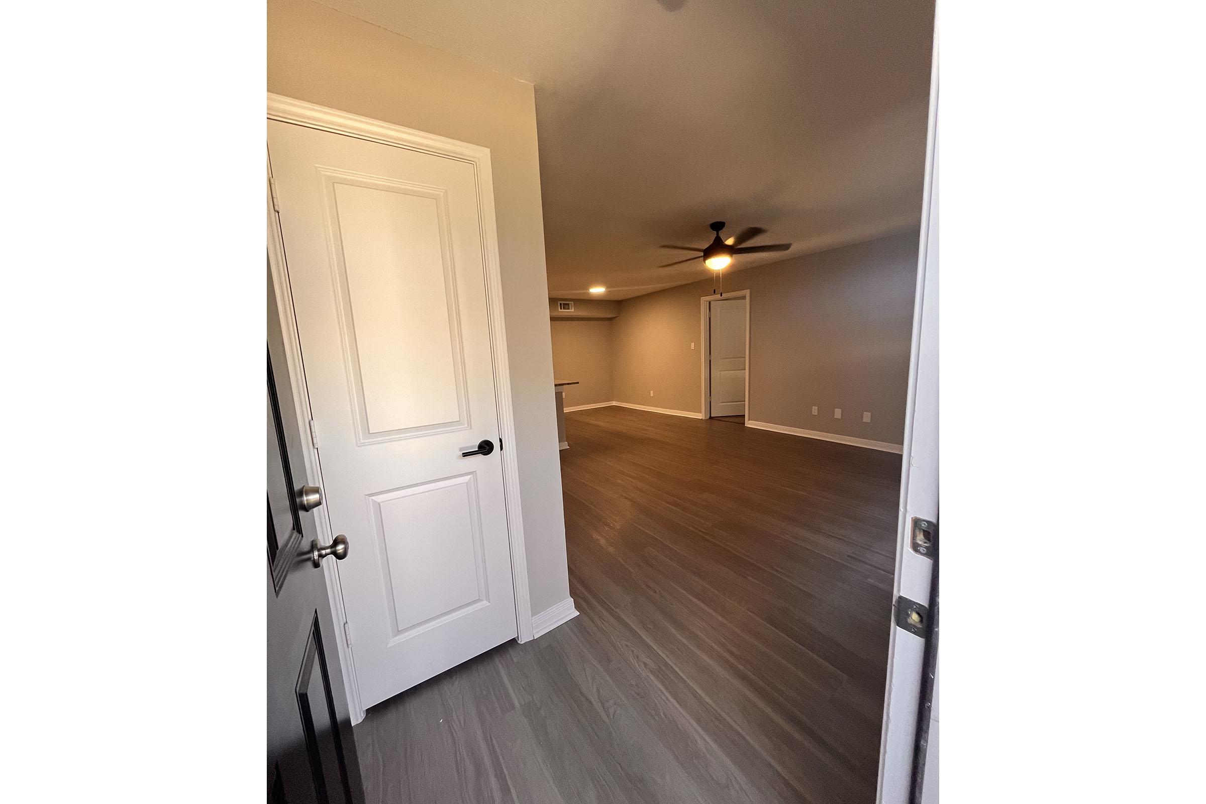 Interior view of a room with gray walls and wooden flooring, featuring a ceiling fan. A door is partially open, leading into the room. There is a light source in the ceiling, and the space appears to be well-lit and inviting.
