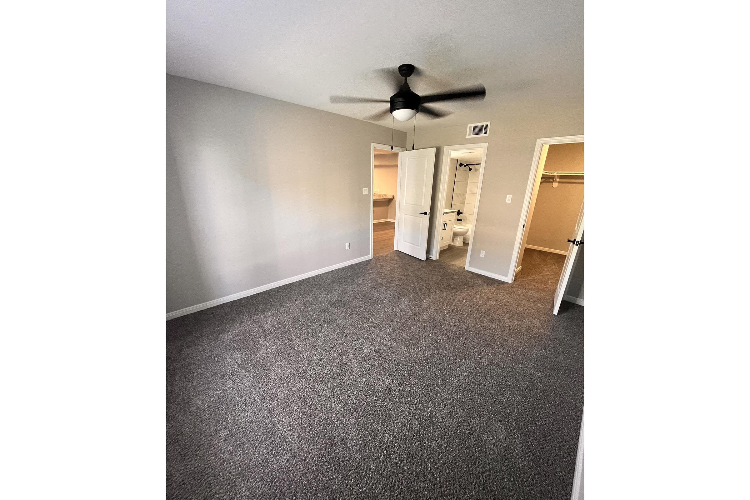 A well-lit, spacious room with gray carpet and light-colored walls. A ceiling fan is installed, and there are two open doors leading to a closet and a bathroom. The room appears clean and offers a neutral and inviting atmosphere, suitable for various uses.