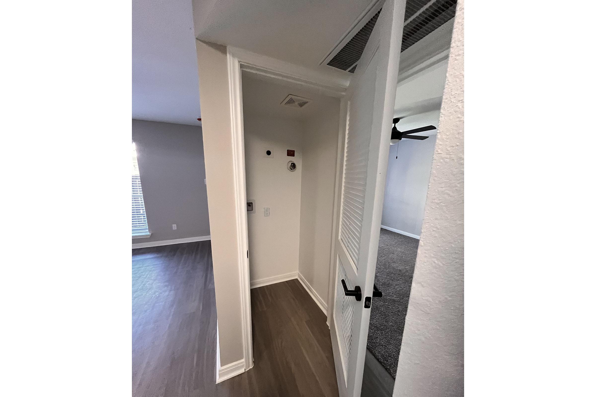 A narrow hallway leading to a room, featuring a partially open door on the left. The wall is painted light color, with a vent above. The flooring is light wood, and at the end of the hallway, there's a carpeted area visible. Sunlight filters in through a window in the adjacent room, enhancing the bright and airy atmosphere.