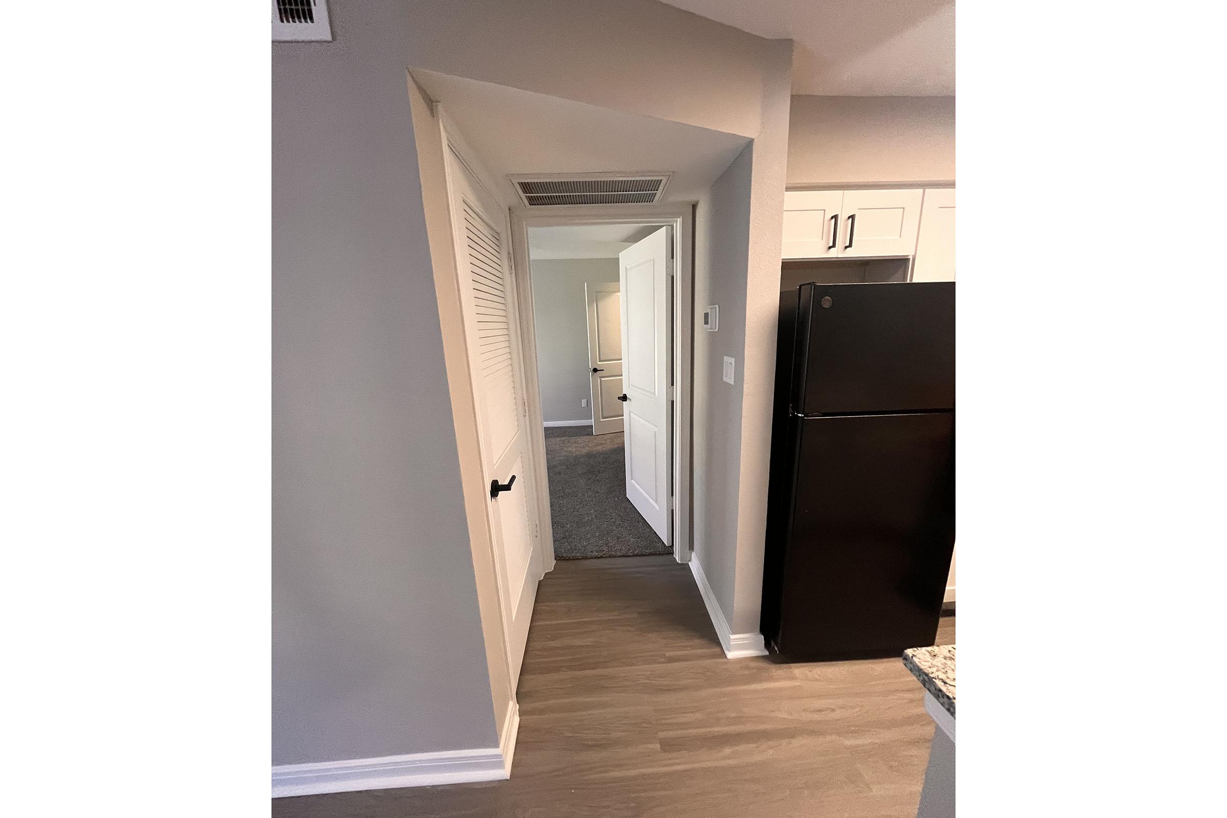A view of a small hallway leading to another room, with a light-colored wall and a doorway on the right. The entrance is framed by a partially open door. A black refrigerator is visible on the left, and the floor features light wood-like tiles.