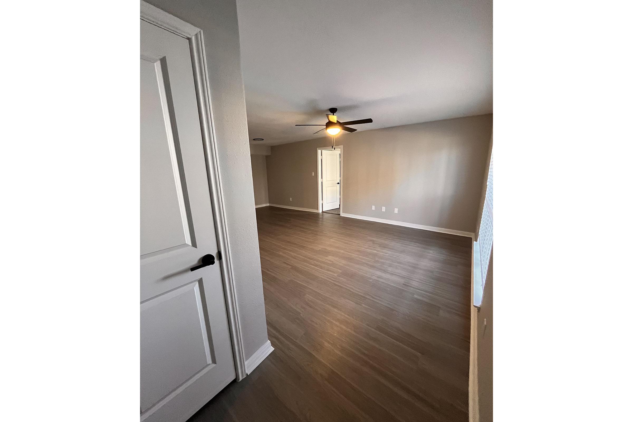 A spacious room with a high ceiling, featuring a ceiling fan and light fixture. The walls are painted in a neutral shade, and the floor is covered with light-colored laminated flooring. A closed door is visible on the left, and the layout suggests an open space leading to another room in the background.