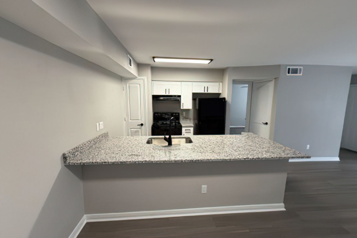 Modern kitchen featuring a granite countertop with a sink, two black appliances (oven and refrigerator), white cabinetry, and open layout. The walls are painted light gray, and there is a recessed ceiling light. The flooring is sleek and dark, complementing the contemporary design.