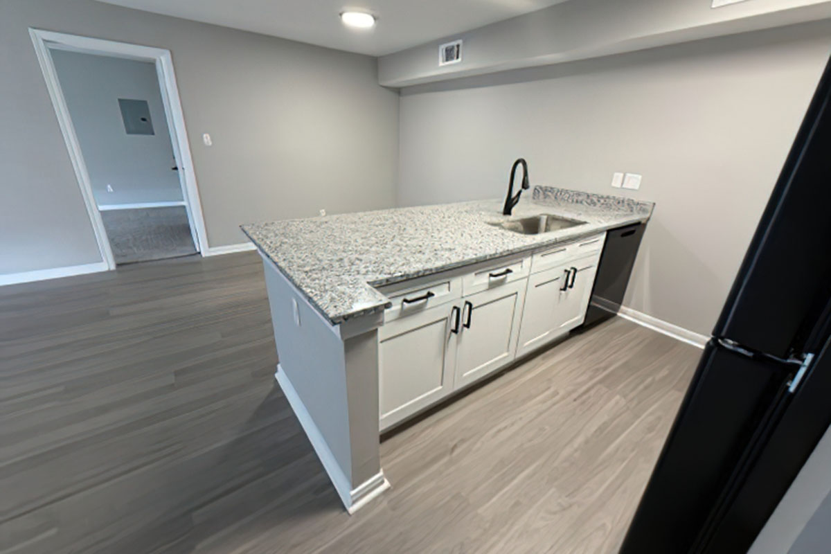 A modern kitchen featuring a granite countertop with a black faucet and sink, white cabinets, and a stainless steel dishwasher. The flooring is a light wood finish, and the walls are painted in a neutral gray tone. A doorway leads to another room in the background.
