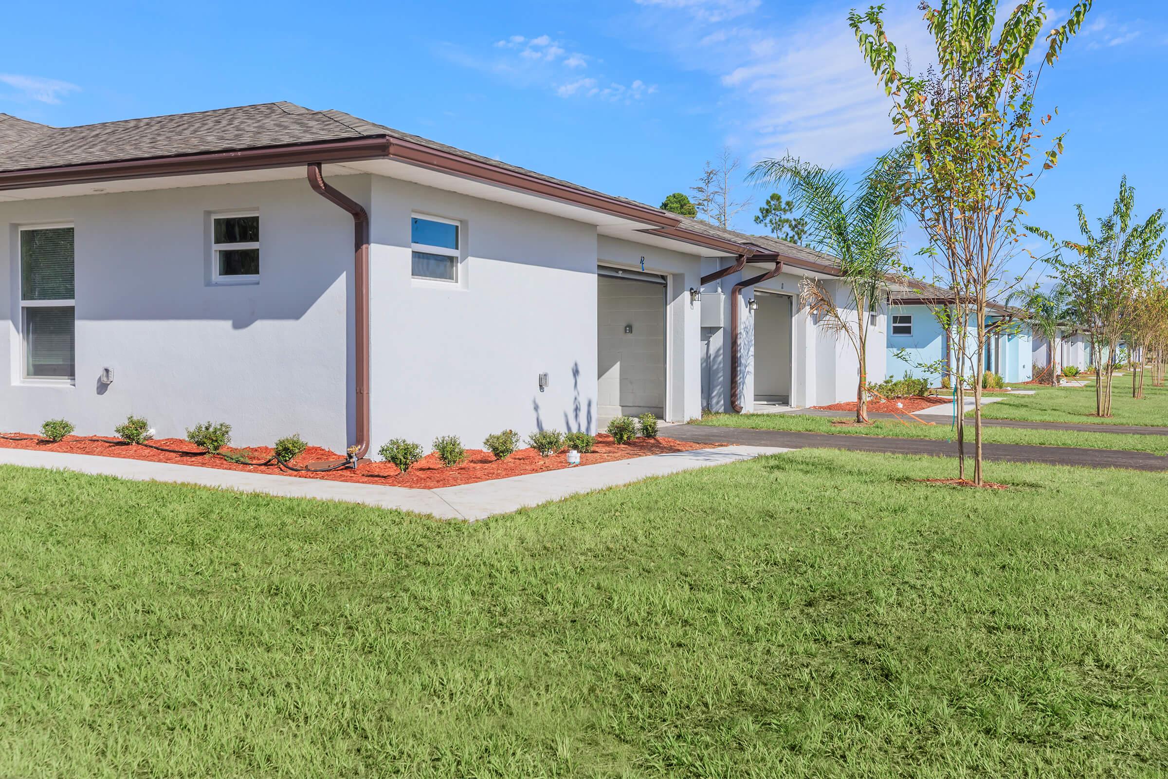 A modern single-story residential building with a light-colored exterior, featuring a driveway and neatly landscaped front yard. The lawn is lush and green, complemented by small shrubs and mulch beds. Clear blue skies are visible above, creating a bright and inviting overall atmosphere.