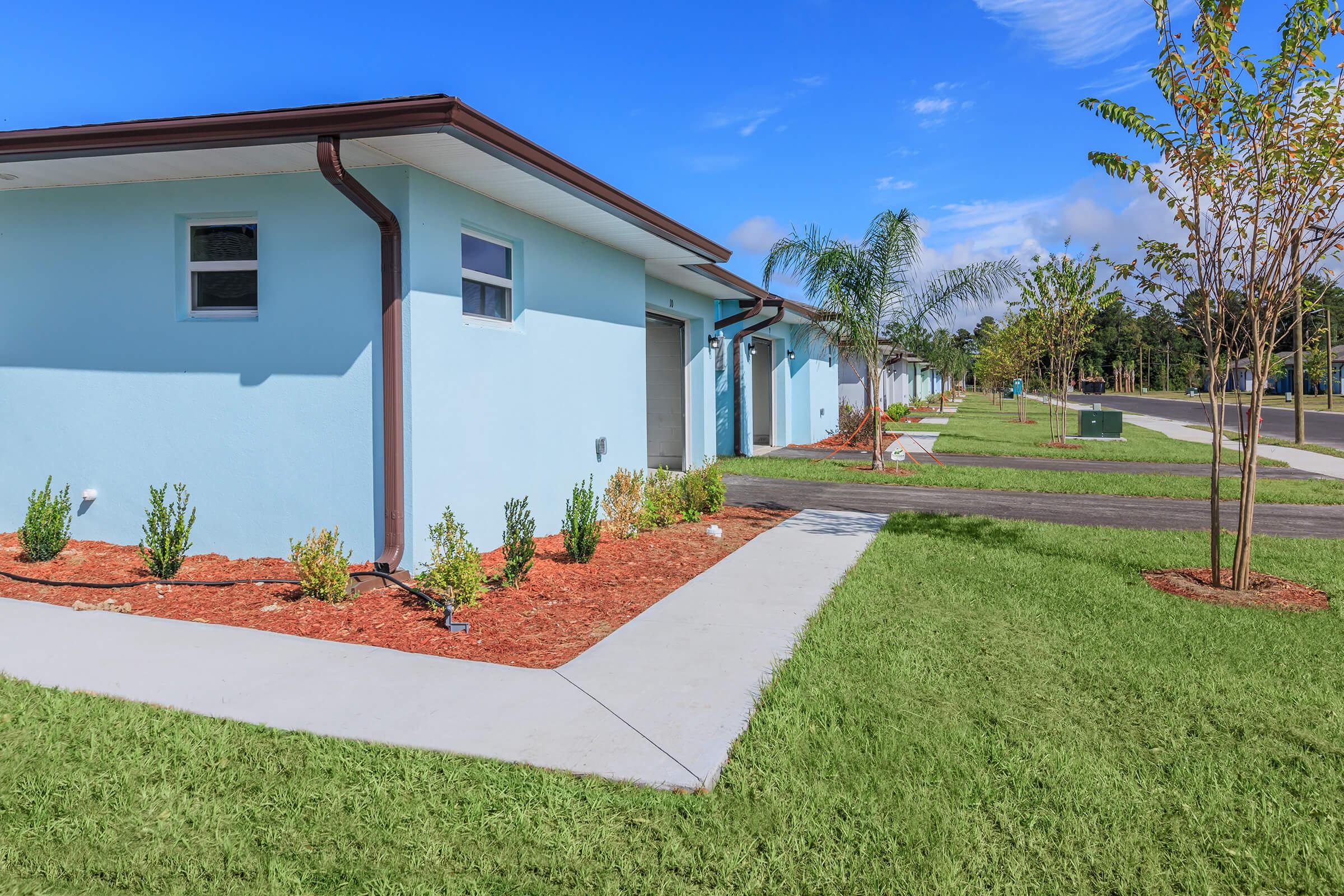 A modern, light blue residential building with a flat roof, featuring a well-manicured lawn and landscaped gardens. Neatly trimmed shrubs and small trees line the pathway, which leads to a paved road. The sky is clear with a few clouds, creating a bright and inviting atmosphere.